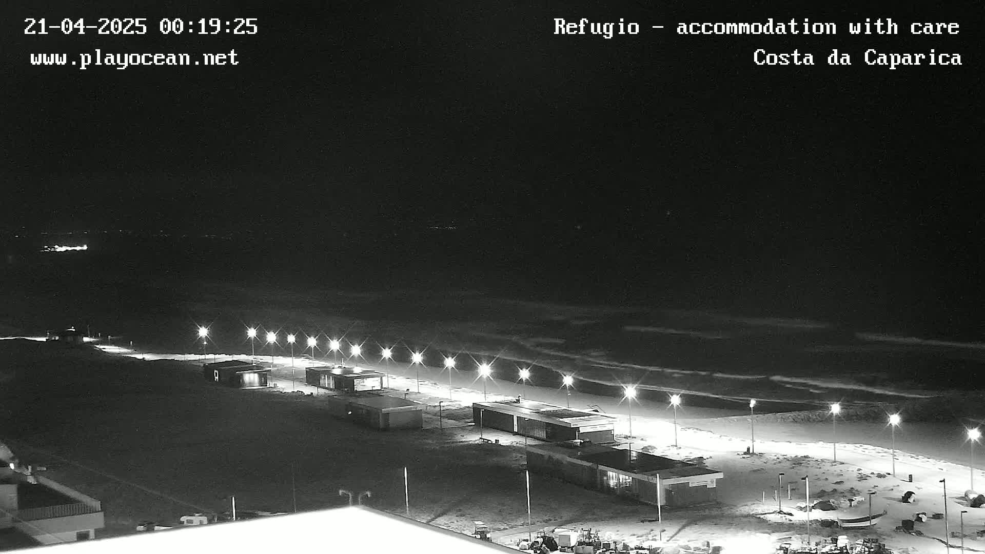 A nighttime, black and white view of a beach at night with gently rolling waves, illuminated by streetlights lining a boardwalk with small buildings.