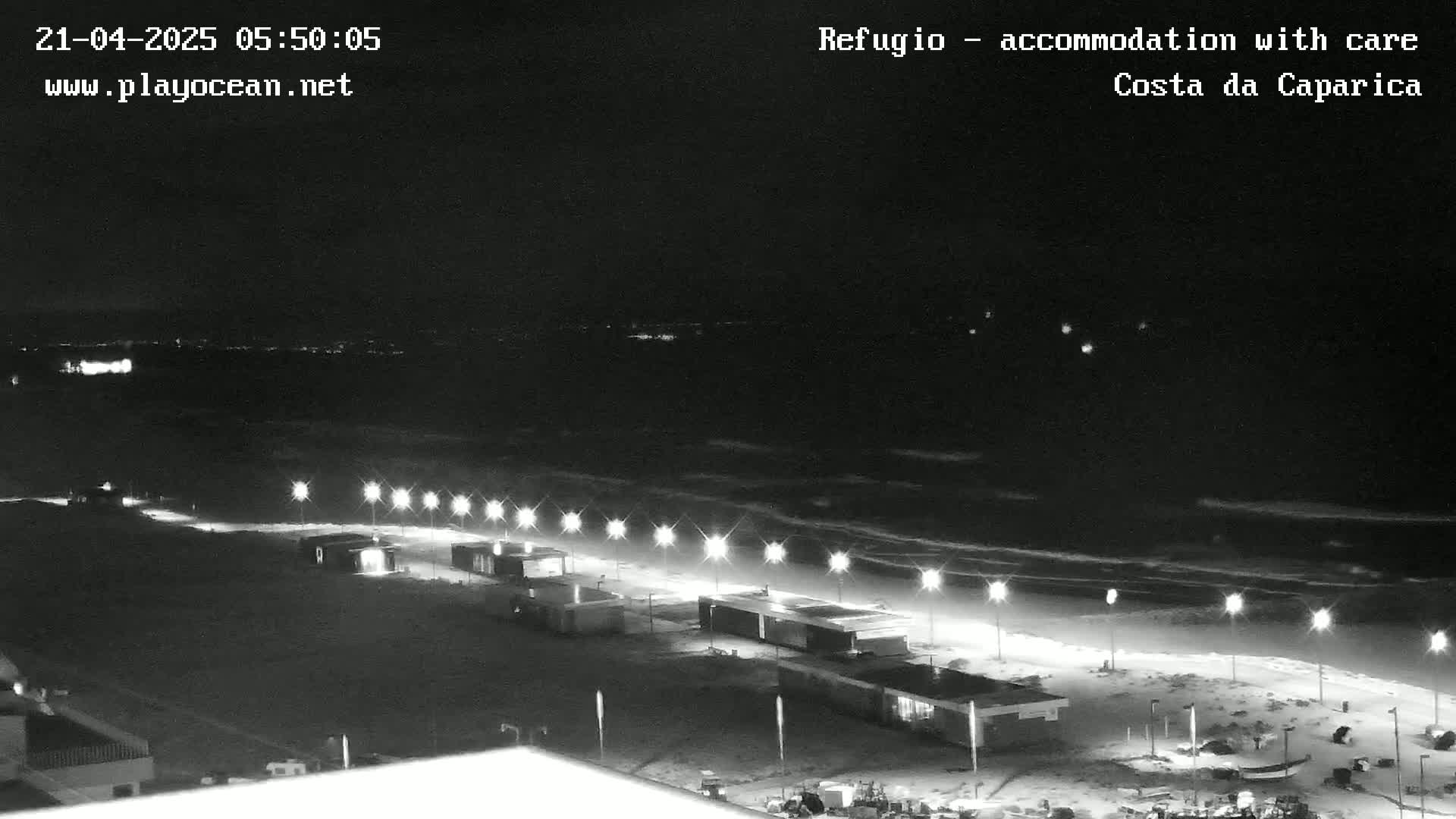 A nighttime beach scene shows a dark ocean with gentle waves lapping at a shoreline lined with illuminated structures and lampposts.