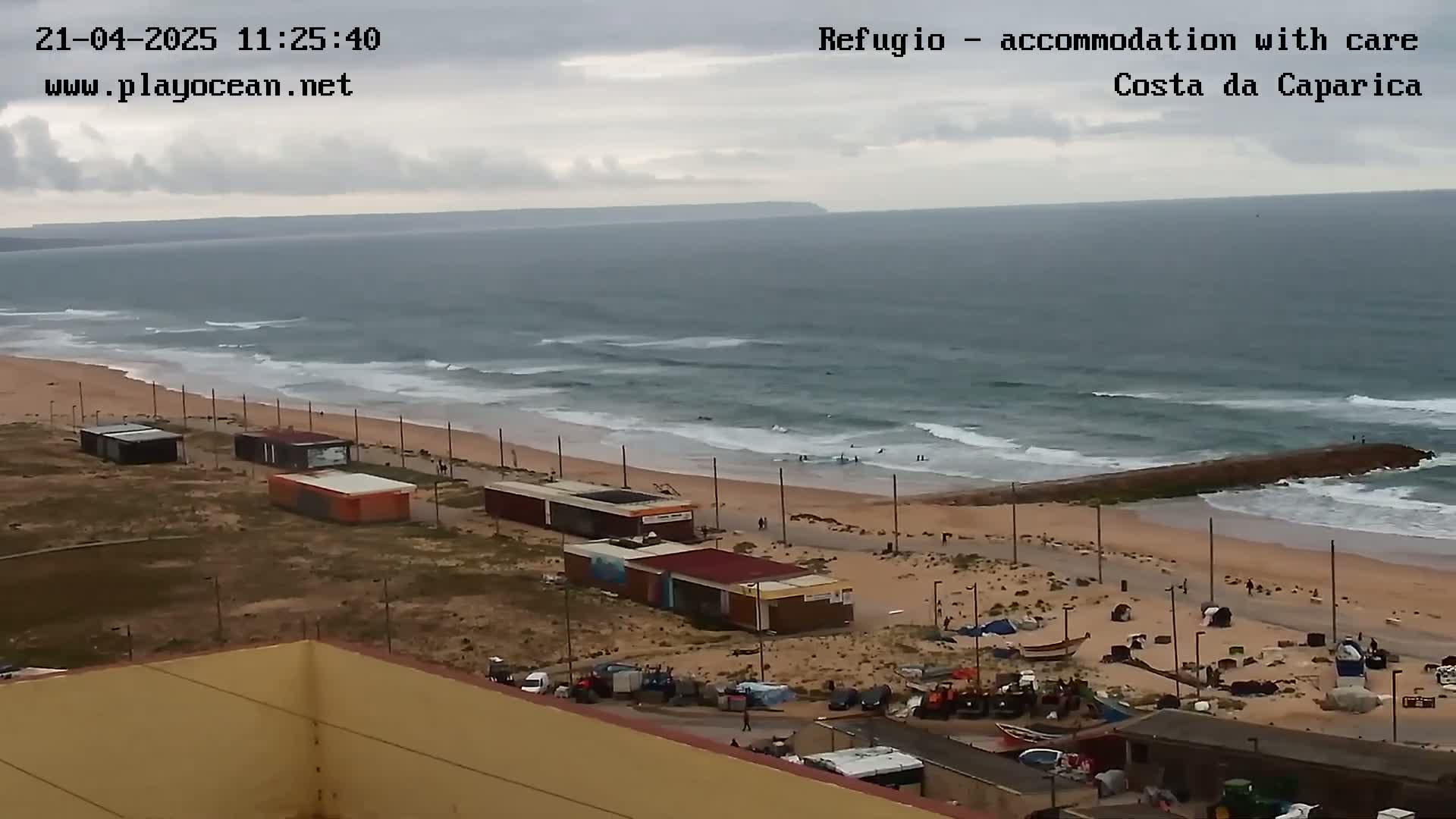 An overcast day shows a beach with small buildings, a rocky jetty, and moderate waves rolling onto the shore.