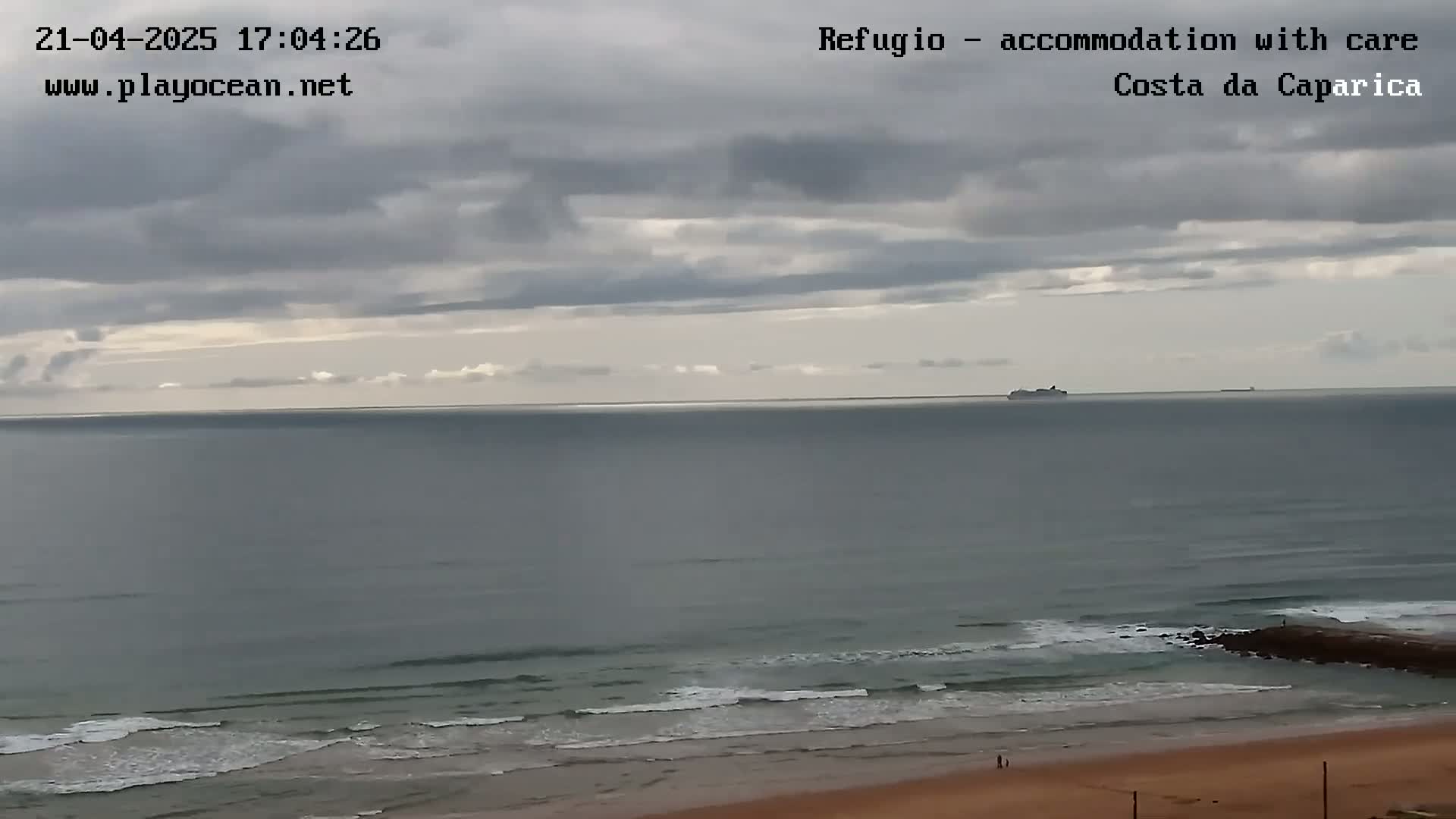 A distant cruise ship is visible on a calm ocean under a cloudy sky, with a sandy beach and small waves in the foreground.