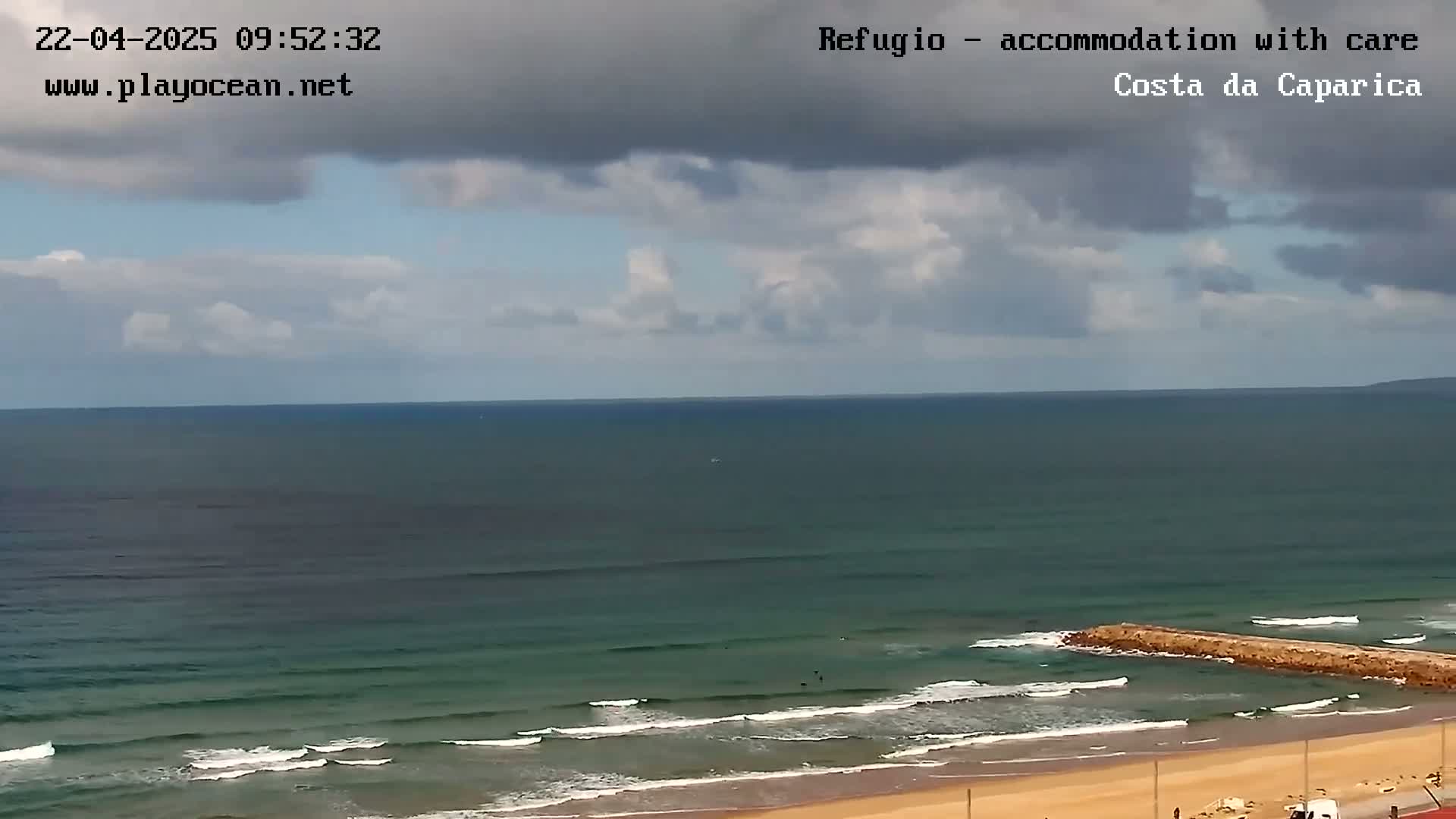 A mostly cloudy sky overlooks a sandy beach with a long rock jetty extending into a dark teal ocean with small waves.