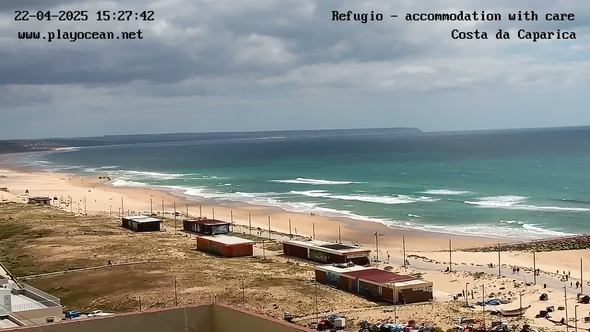 A sandy beach with small buildings and a few people stretches along a turquoise ocean under a partly cloudy sky.