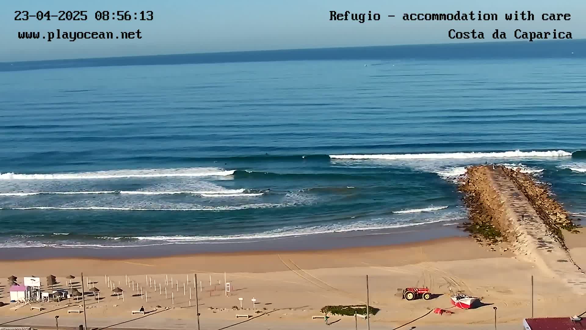 A sandy beach with several surfers in the ocean under a clear sky, a rock jetty extending into the water, and some beach equipment and vehicles on the sand.