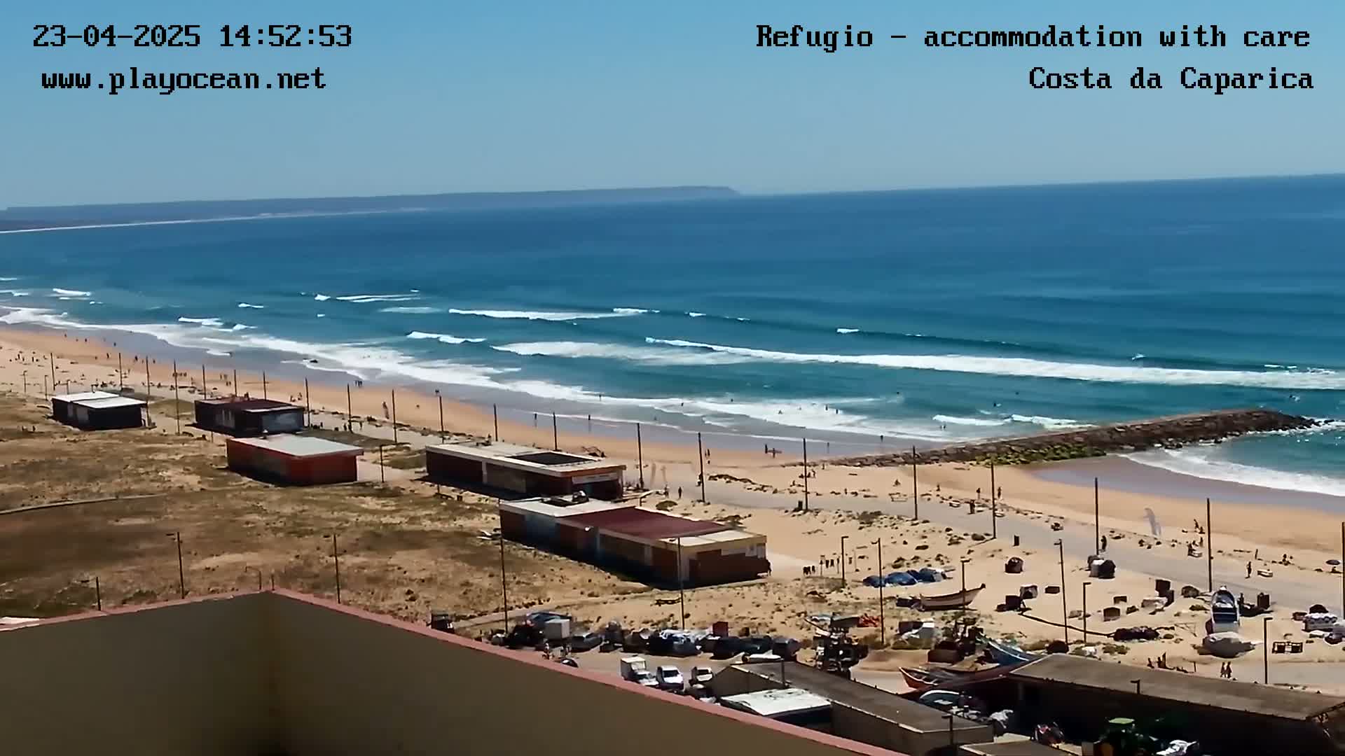 A sandy beach with small buildings and a parking lot is shown, overlooking a wide ocean with rolling waves under a clear, sunny sky.