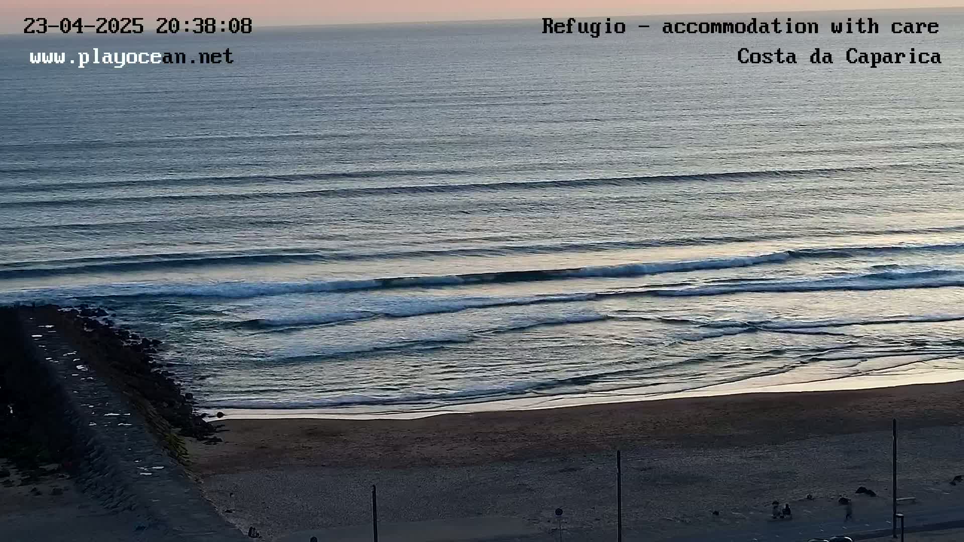 A sandy beach at dusk is seen from an elevated viewpoint, showing gentle waves rolling onto the shore under a twilight sky.