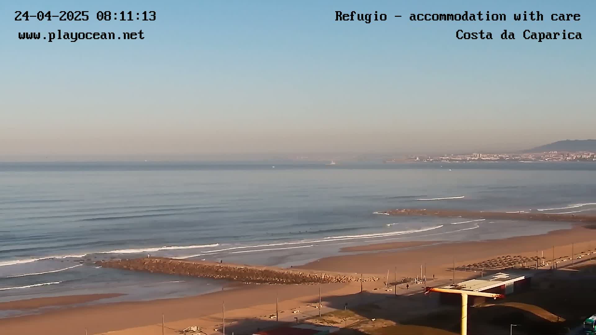 A sandy beach with a calm ocean and a distant cityscape under a clear sky.