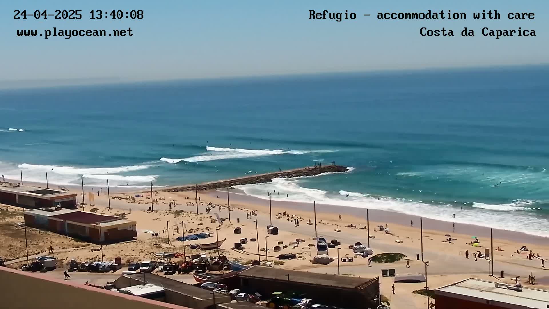 A sandy beach with several people and parked cars is shown, with a rocky jetty extending into a blue ocean with waves breaking.