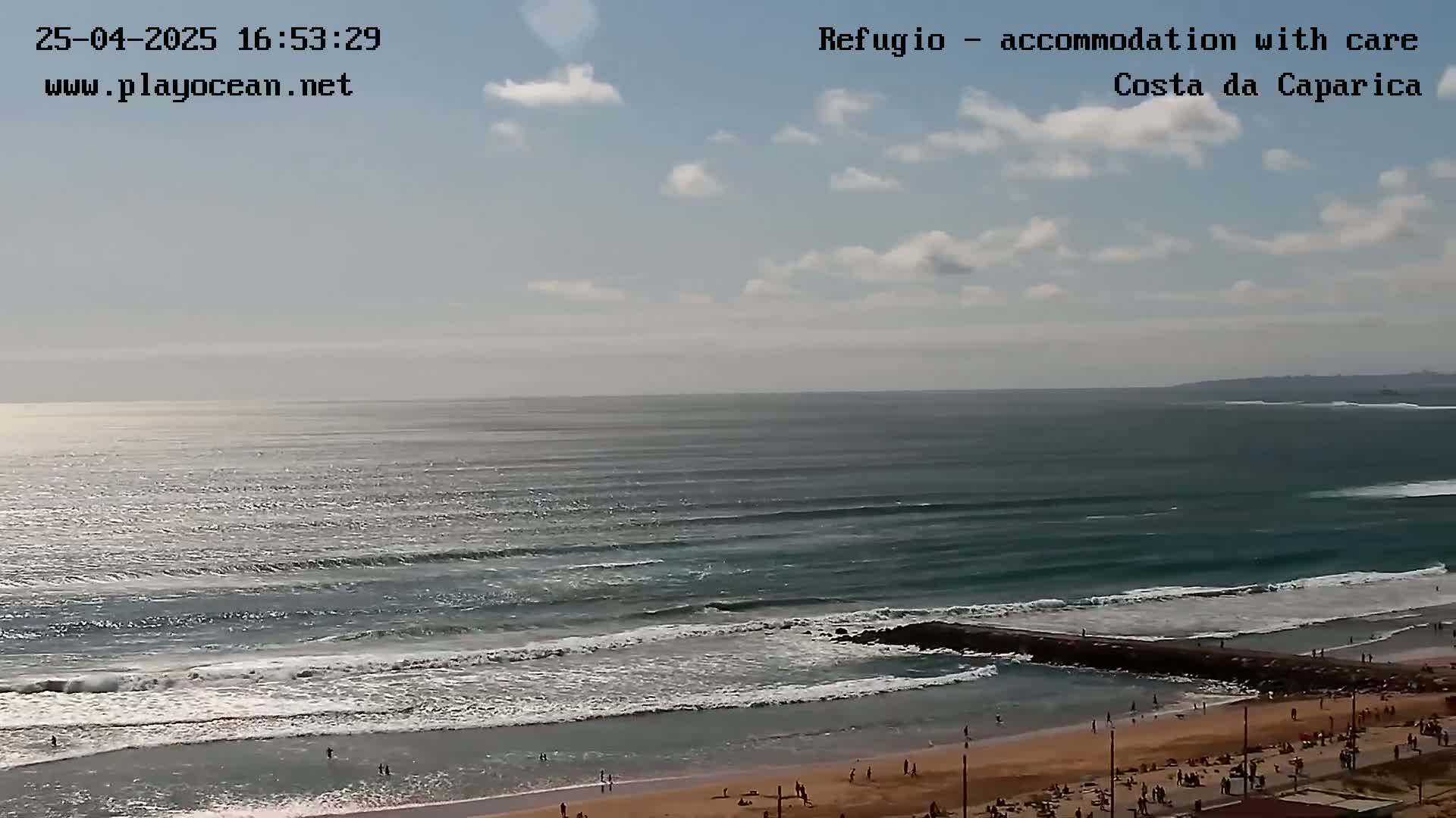 A beach with people sunbathing and swimming, under a partly cloudy sky, shows moderate waves breaking near a jetty.