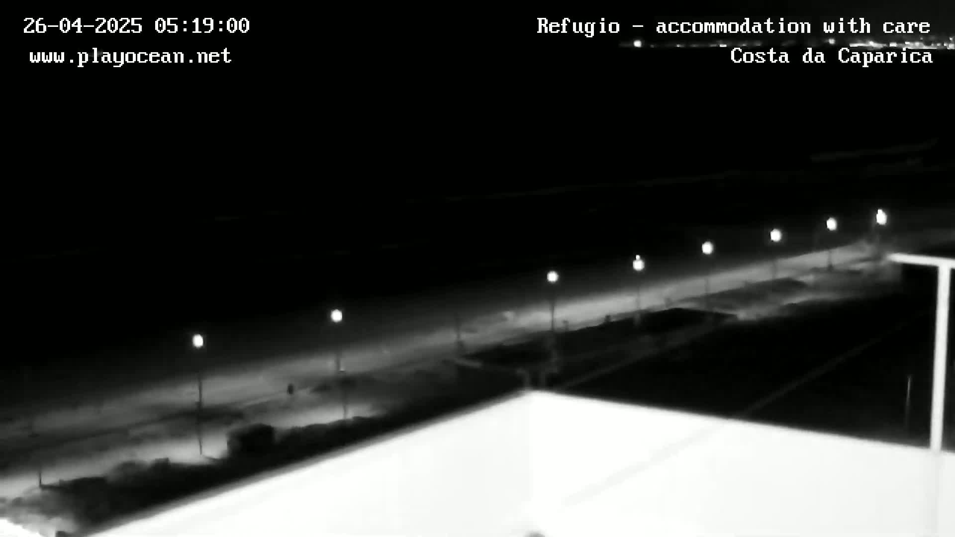 A nighttime, black and white view shows a dark beach with several lights along a walkway, seen from an elevated perspective.