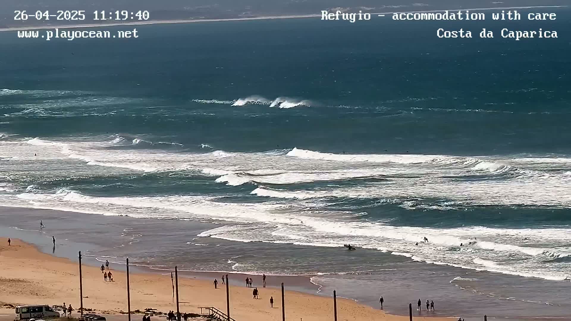 A wide sandy beach with people walking along the shore and several surfers in the water, under a partly cloudy sky, with waves rolling in from a deep blue ocean.