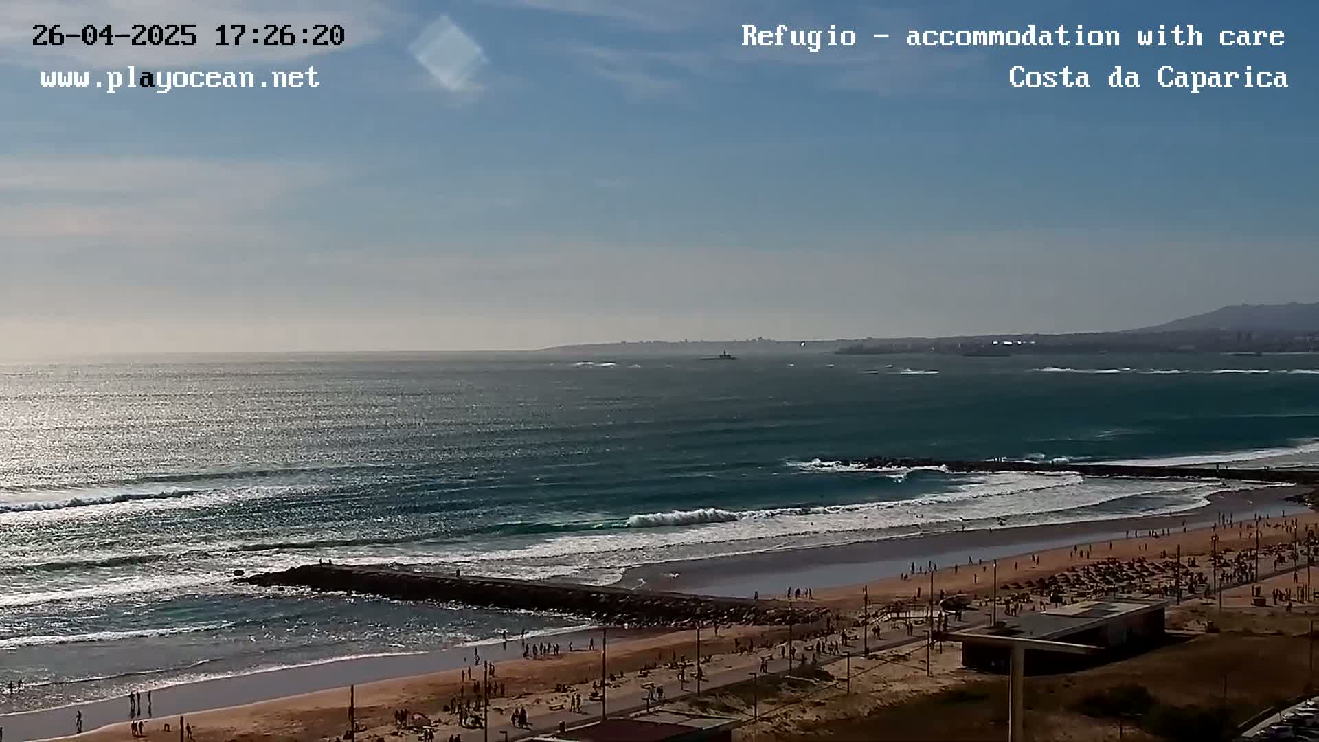 A mostly sunny day shows a long expanse of ocean with waves breaking on a sandy beach where people are relaxing and walking along the shore.