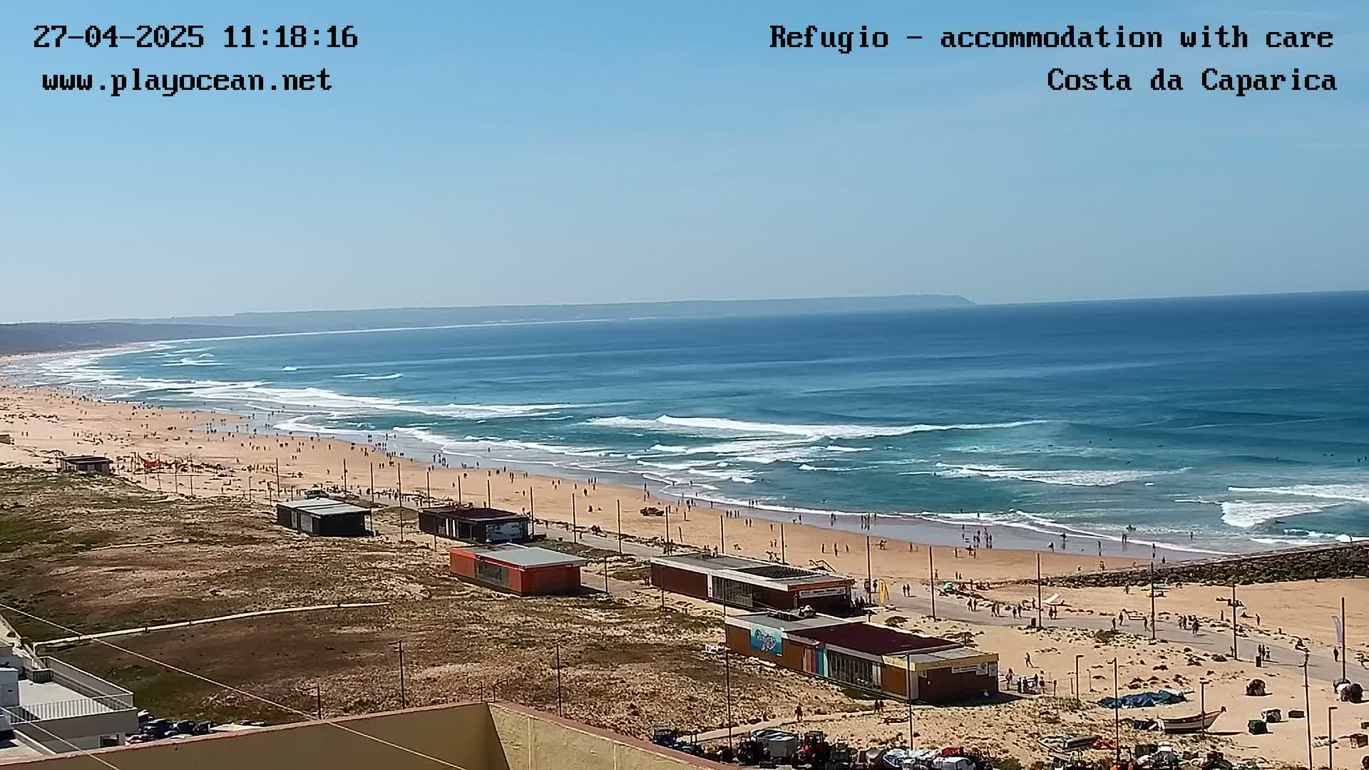 A sunny day at a beach shows a long stretch of sandy shore with people and small buildings, and ocean waves rolling in under a clear blue sky.