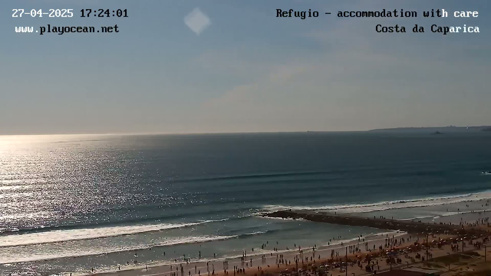 A sunny day at a beach shows a vast ocean with gentle waves breaking on the shore, where many people are enjoying the sand.