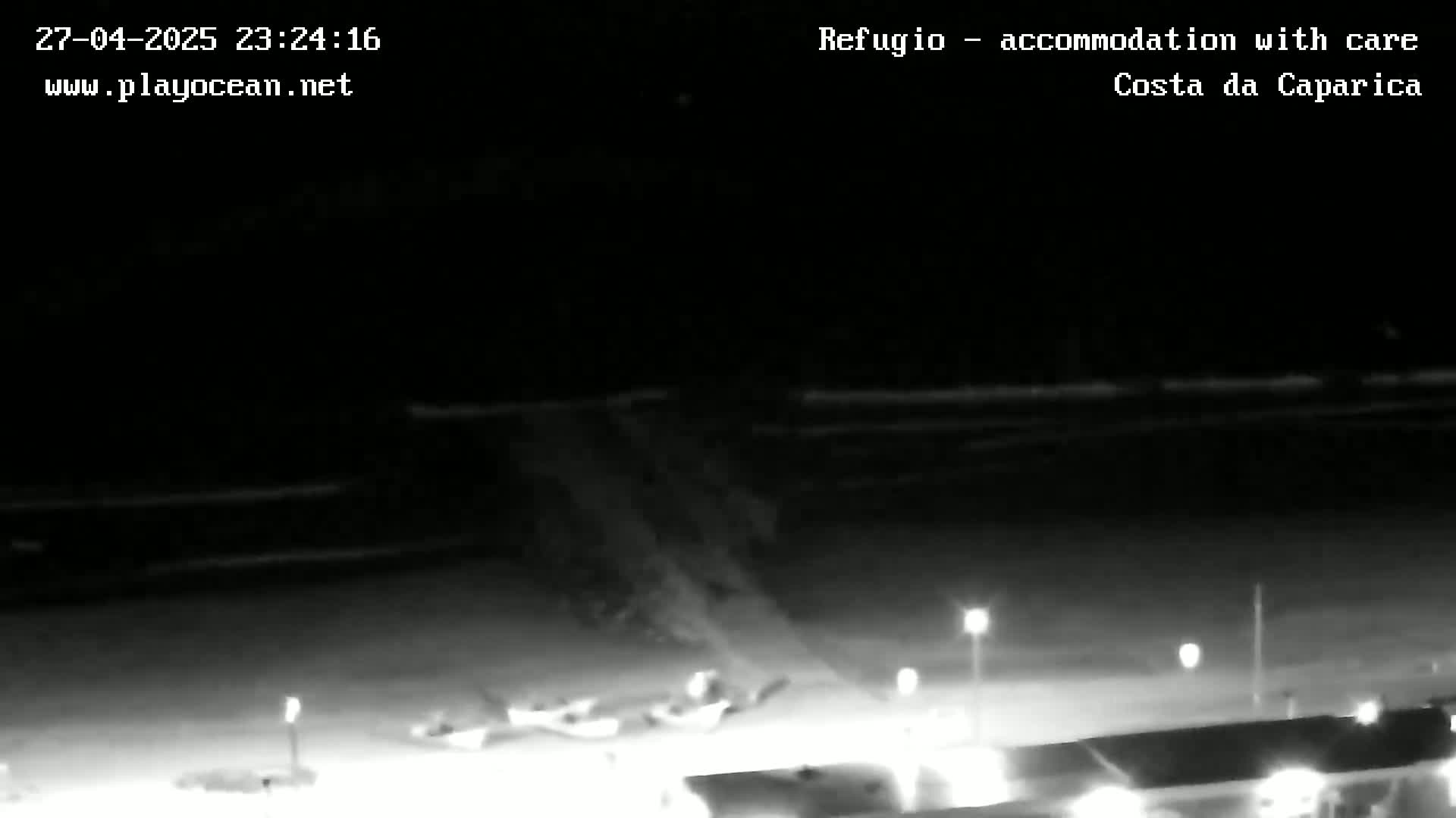 A nighttime, low-light view of a beach area with several small objects near the water and lights illuminating a roadway.