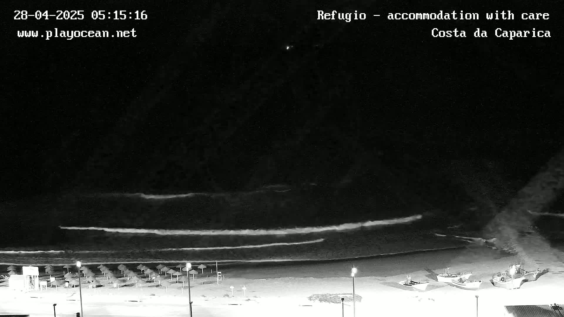 A nighttime, black-and-white view of a beach with several small boats pulled ashore, calm waves lapping the sand, and closed umbrellas lining the shoreline.