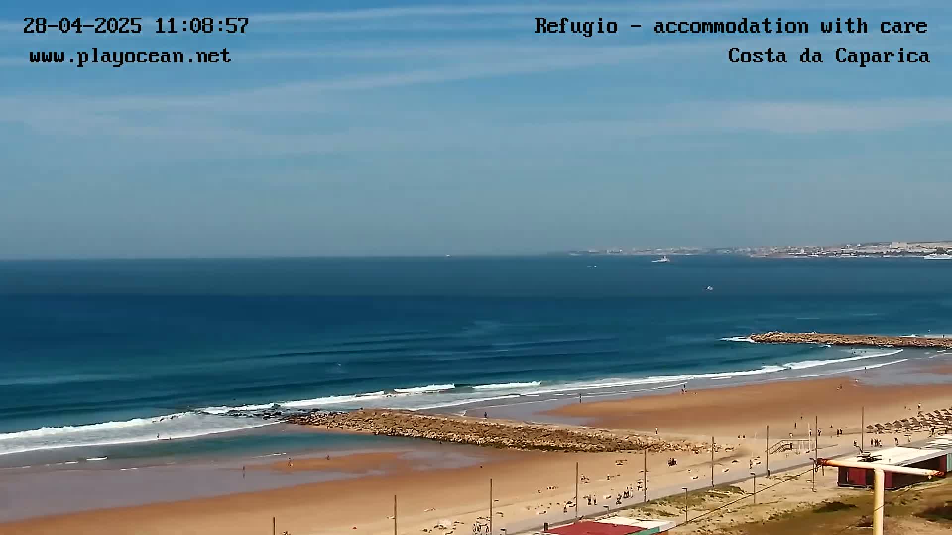 A sandy beach with people sunbathing and swimming, a rock jetty, and rolling ocean waves under a mostly clear sky.