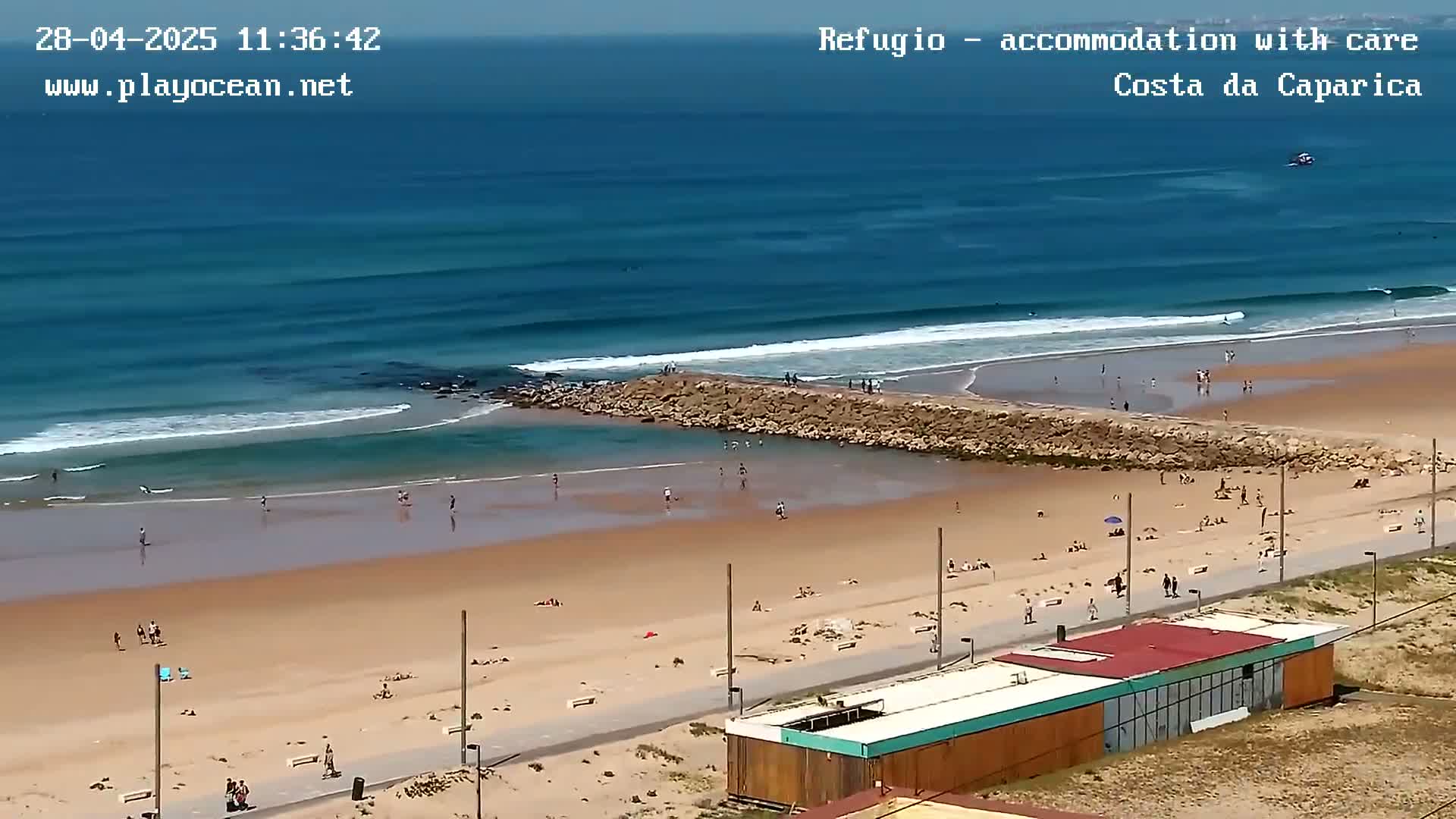 A sandy beach with people swimming and sunbathing, calm ocean waves breaking near a rock jetty under sunny skies.