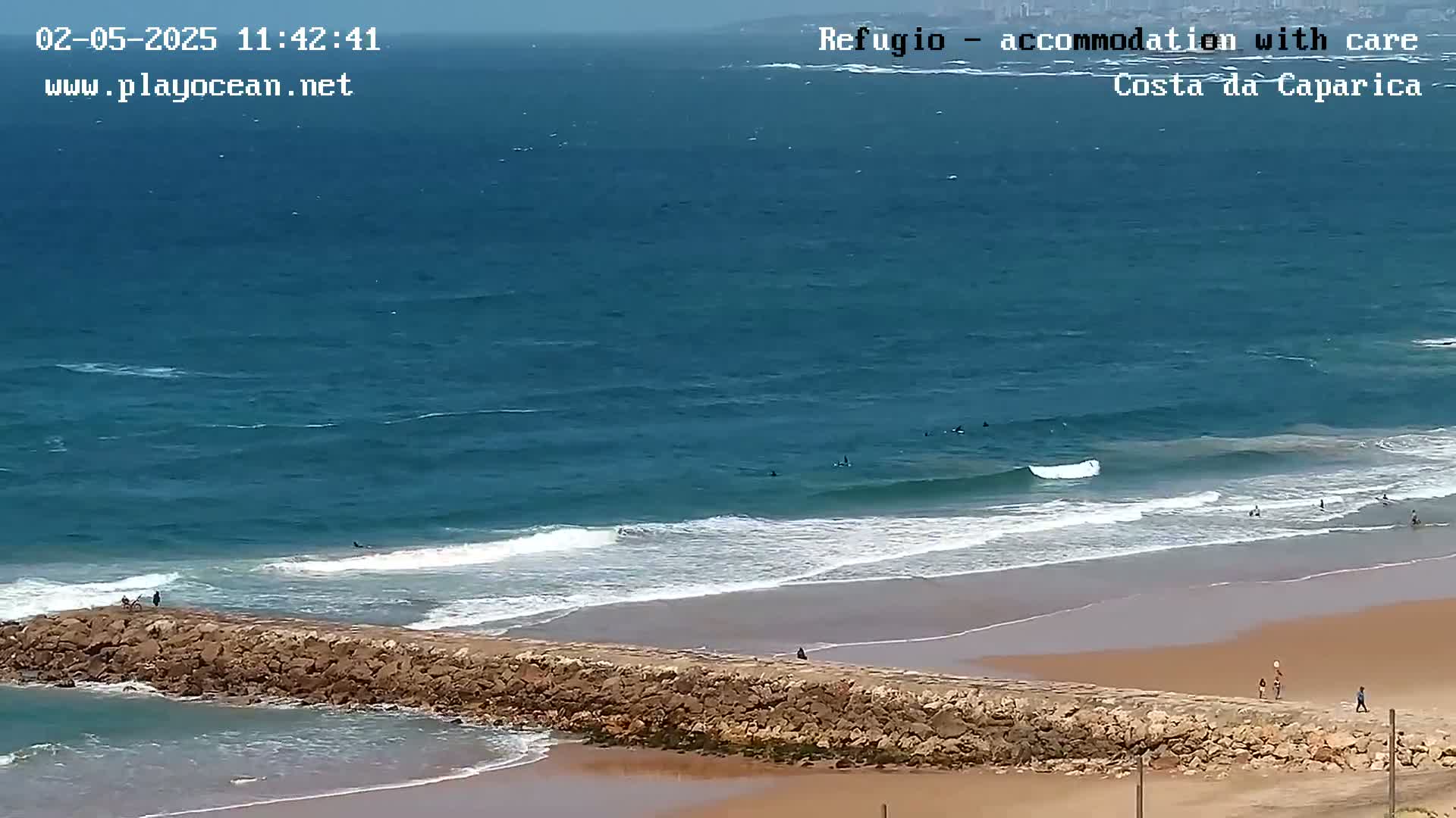 A sandy beach with a long stone wall extending into the ocean, where several people are surfing and swimming in relatively calm, clear water under a mostly clear sky.