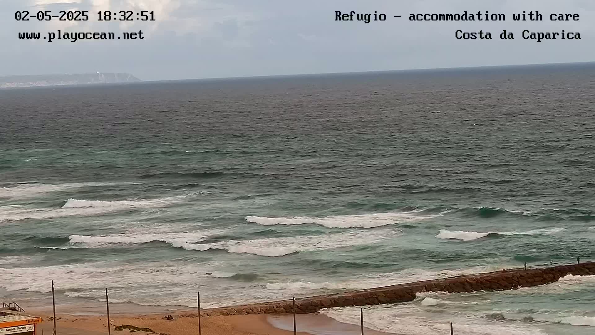 A view of a sandy beach and ocean with moderate waves under an overcast sky shows a distant headland.