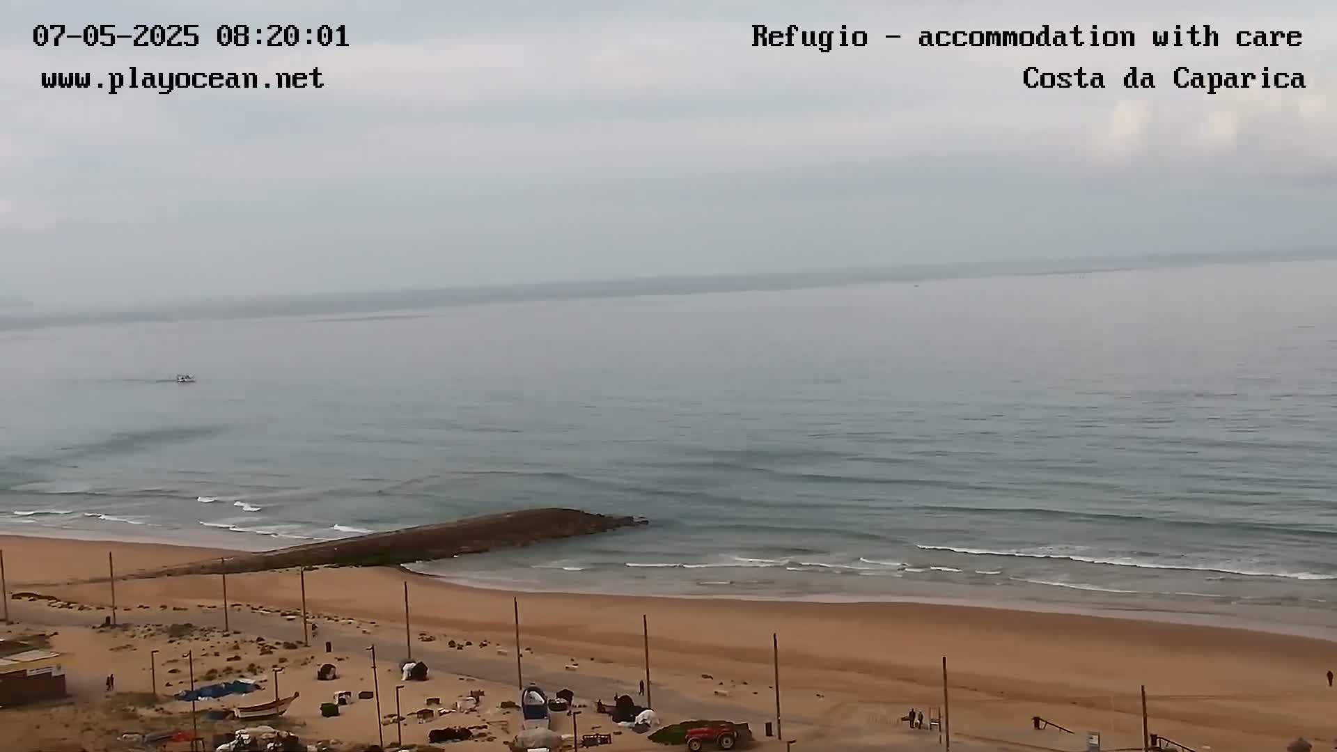 A mostly cloudy day reveals a sandy beach with small waves lapping the shore, a dark structure jutting out into the water near the beach's edge, and several small objects and people near the beach.