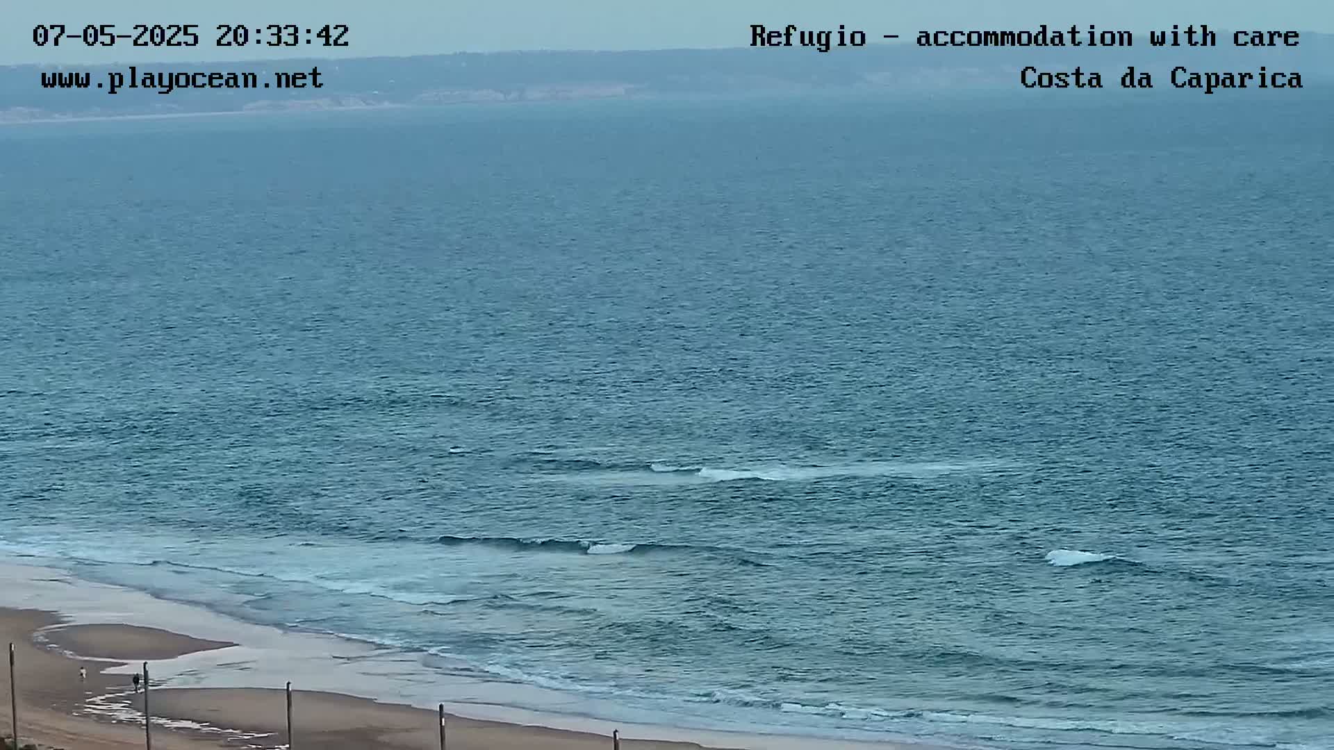 A sandy beach meets a calm, teal ocean with small waves under an overcast sky.