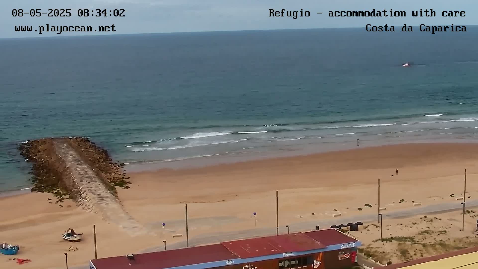A sandy beach with a long rock jetty extends into a calm ocean with small waves under an overcast sky.