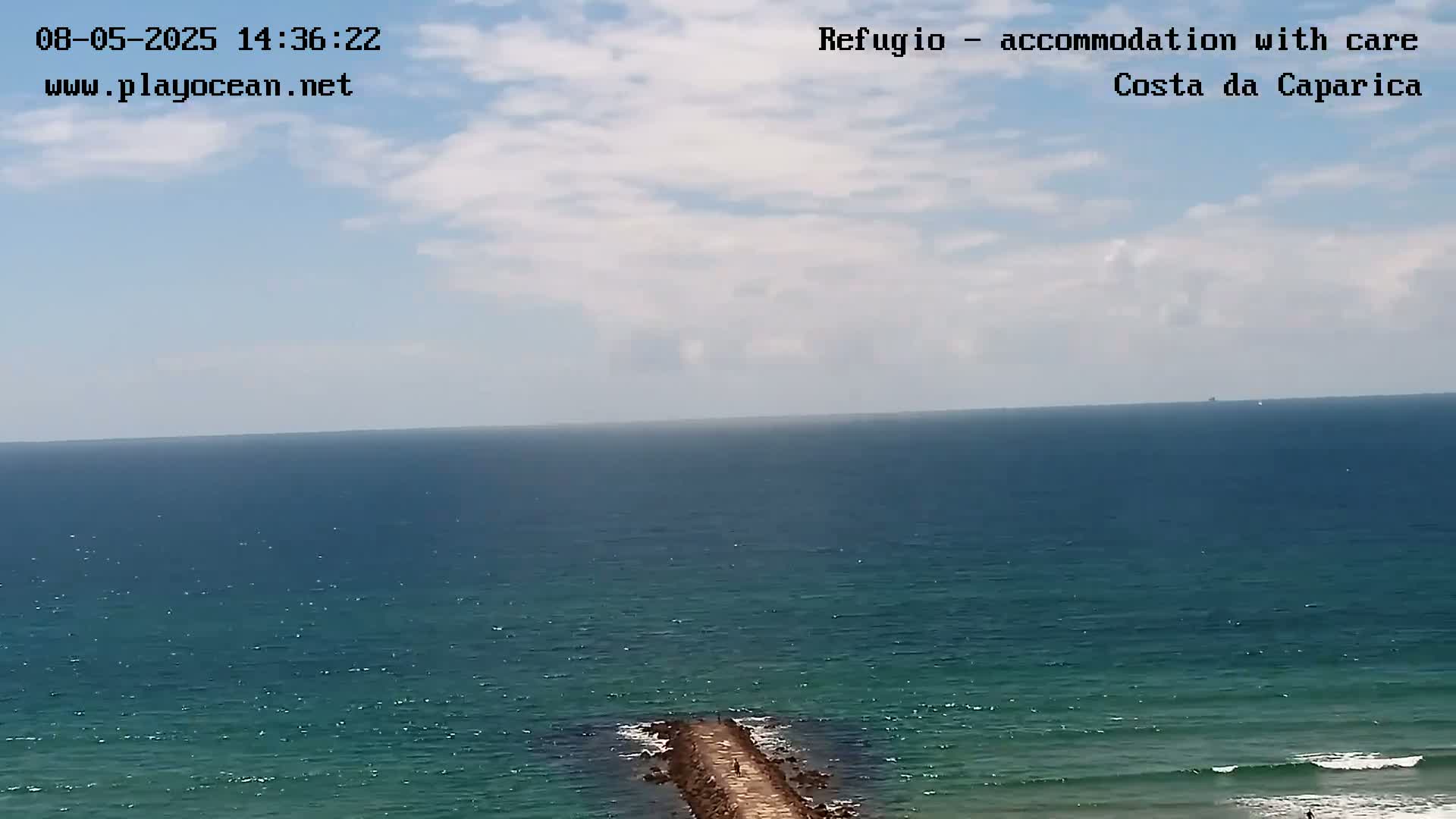 A long, dark-blue ocean stretches to the horizon under a partly cloudy sky, with a stone jetty extending from the shore into the water.