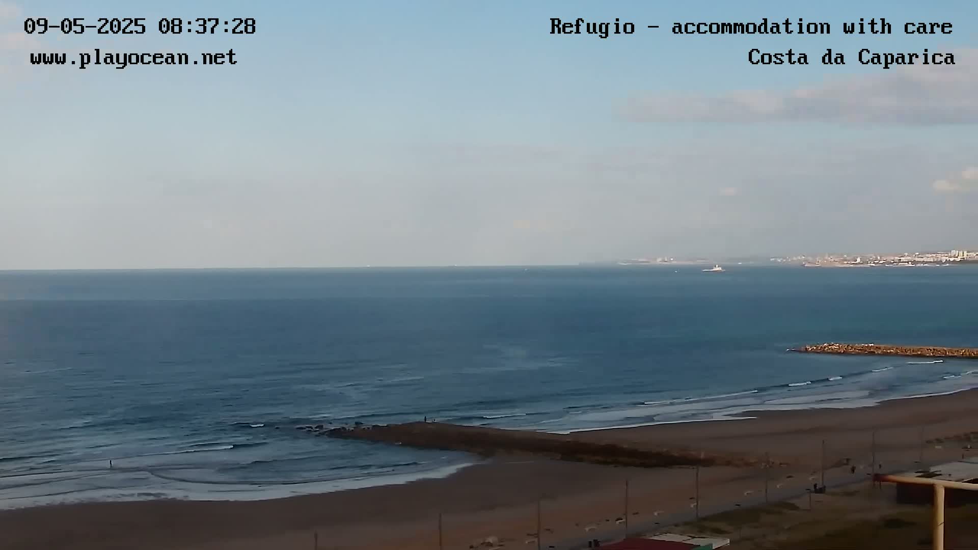 A mostly sunny day reveals a wide sandy beach with gentle waves lapping the shore, a distant city skyline visible across a calm ocean.