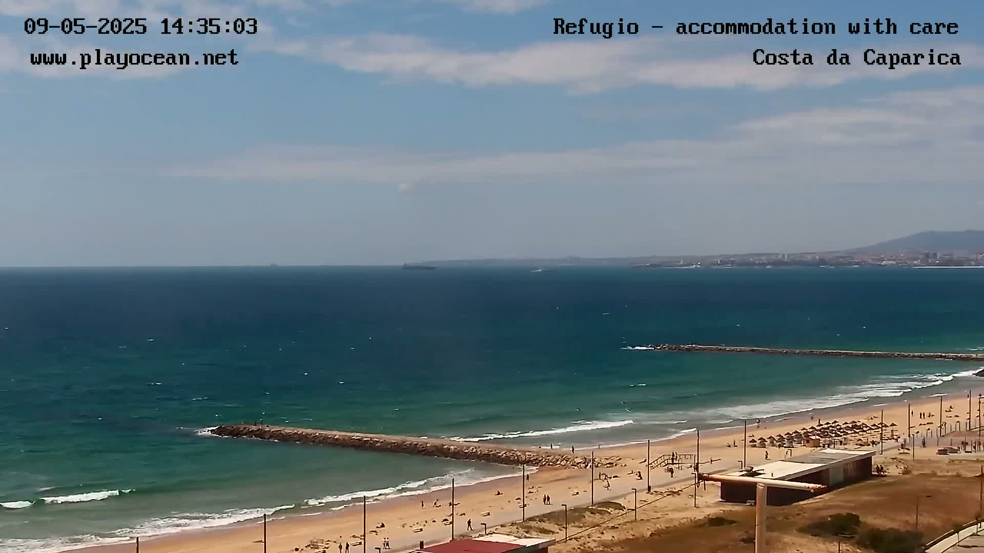 A partly sunny day shows a sandy beach with a few people, some beach umbrellas, and a breakwater extending into a blue ocean with a distant coastline under a partly cloudy sky.