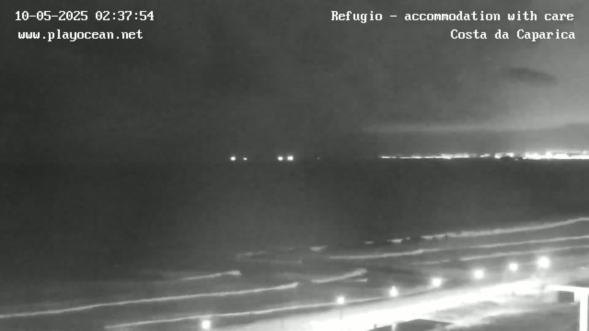 A nighttime, overcast view of a dark ocean with waves lapping a beach, dimly lit by scattered lights along a road.