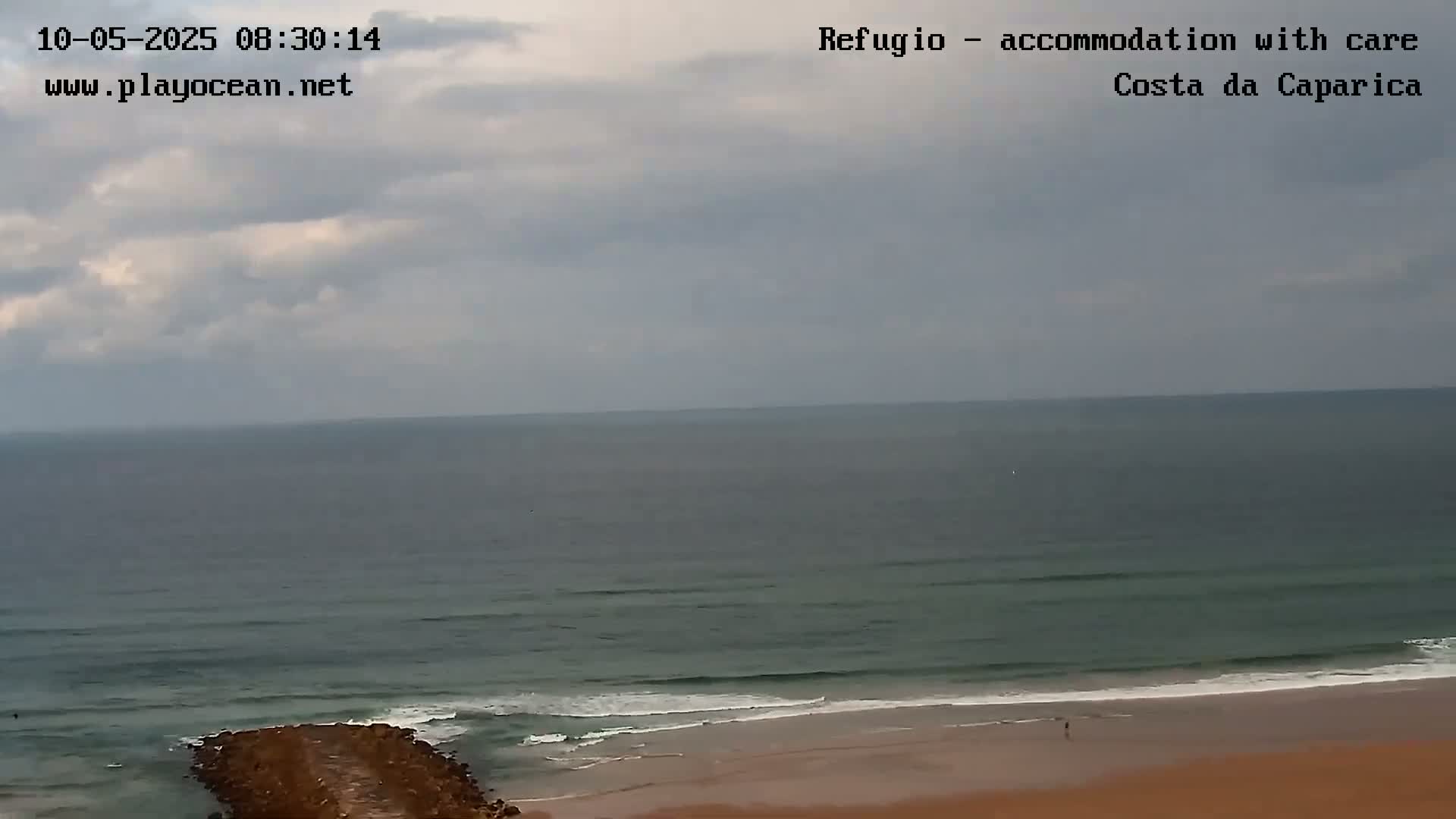 A mostly cloudy sky overlooks a calm ocean meeting a sandy beach with a rock jetty.