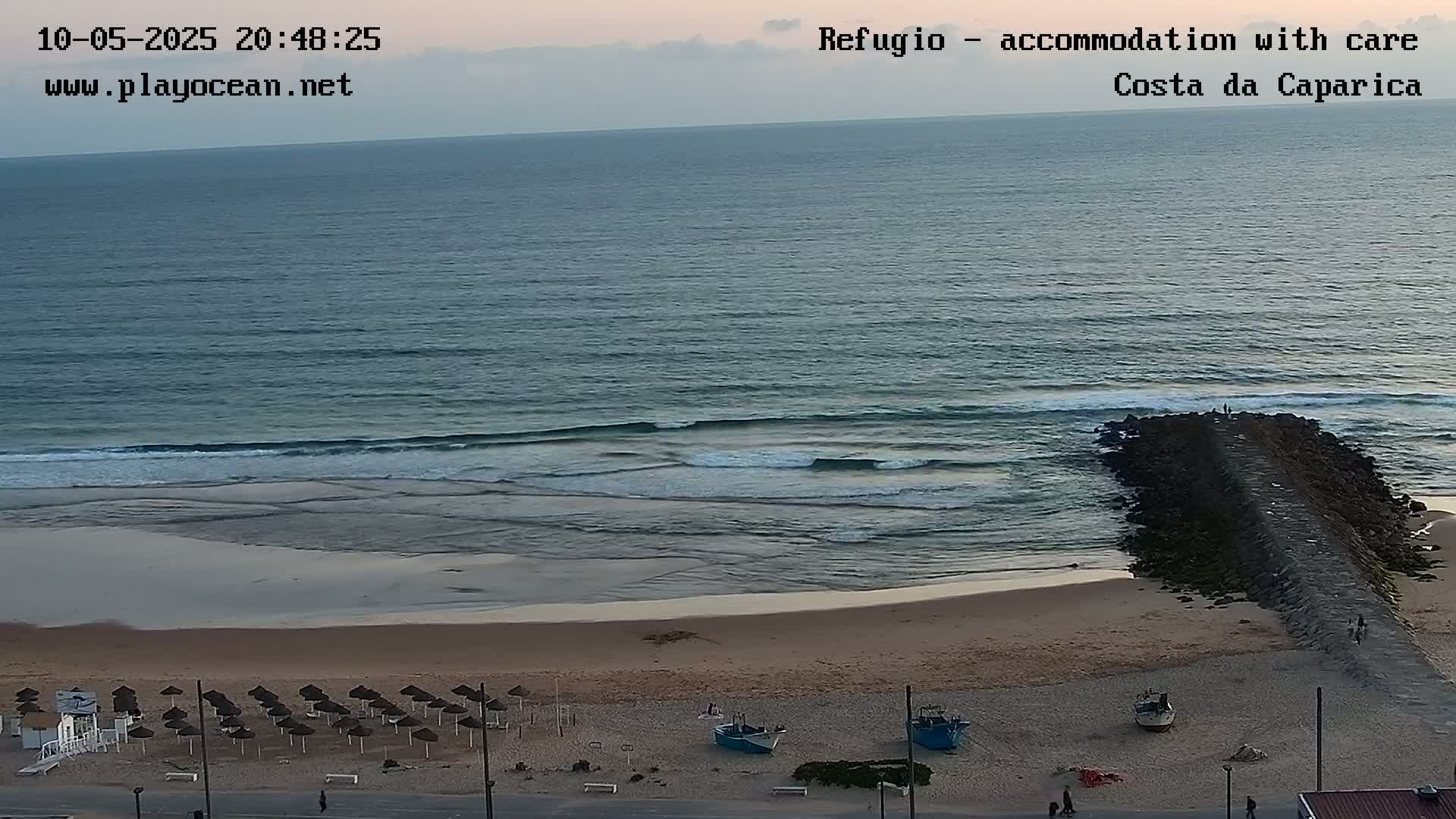 A sandy beach with rows of closed umbrellas and small boats sits next to a calm ocean with gentle waves under a twilight sky.