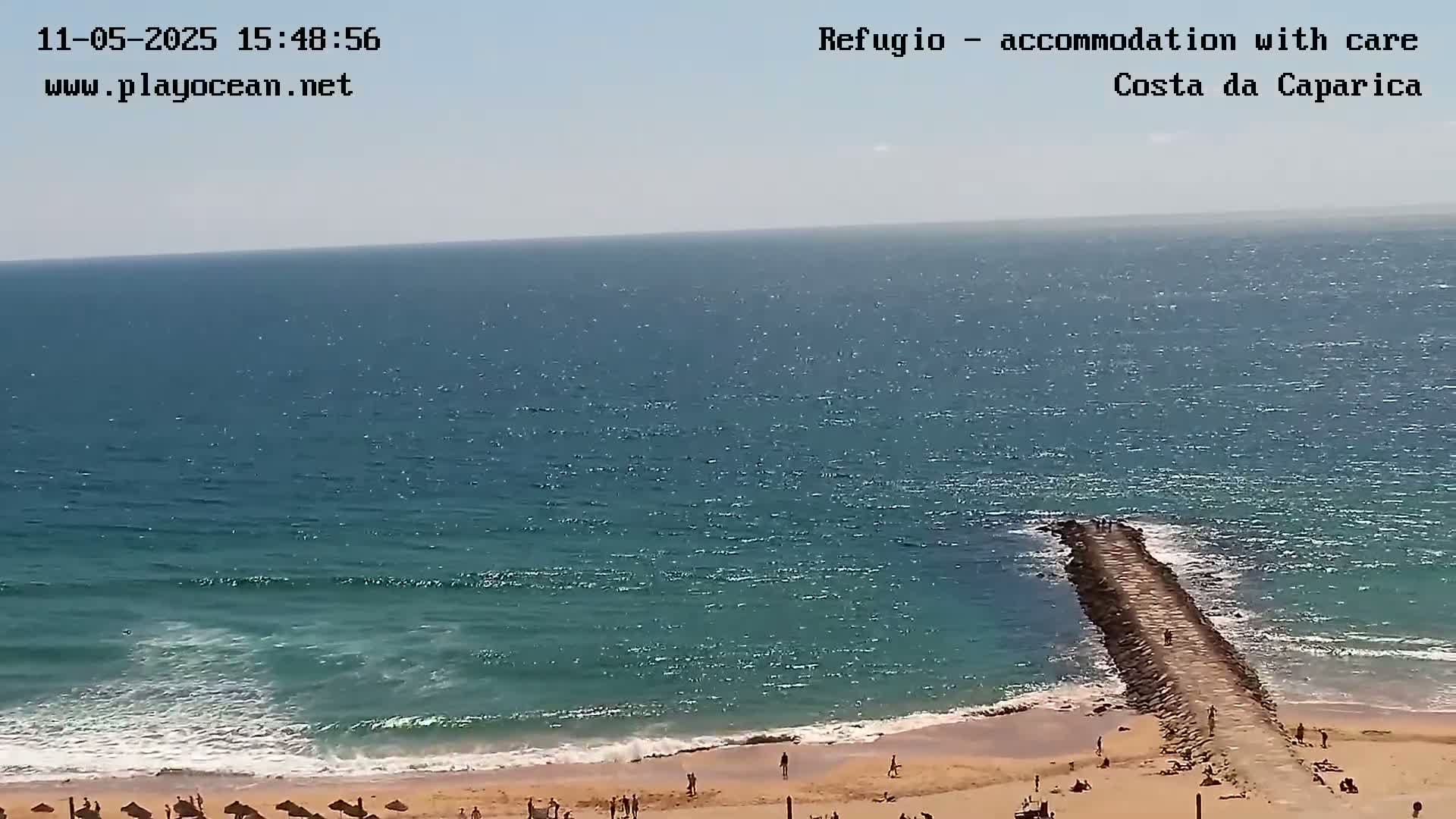 A sandy beach with people on it is shown, along with a long rock jetty extending into a teal ocean on a sunny day with light waves.