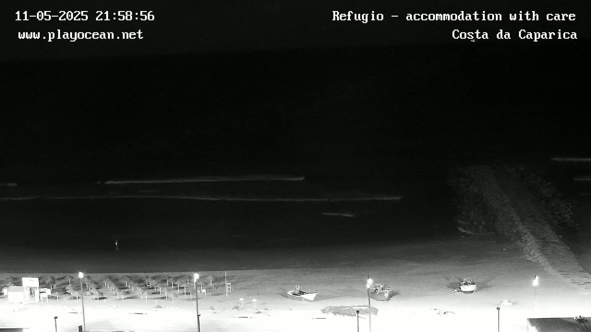 A nighttime beach scene shows a dark ocean with small waves, a mostly empty beach with a few small boats pulled ashore, and rows of closed beach umbrellas.