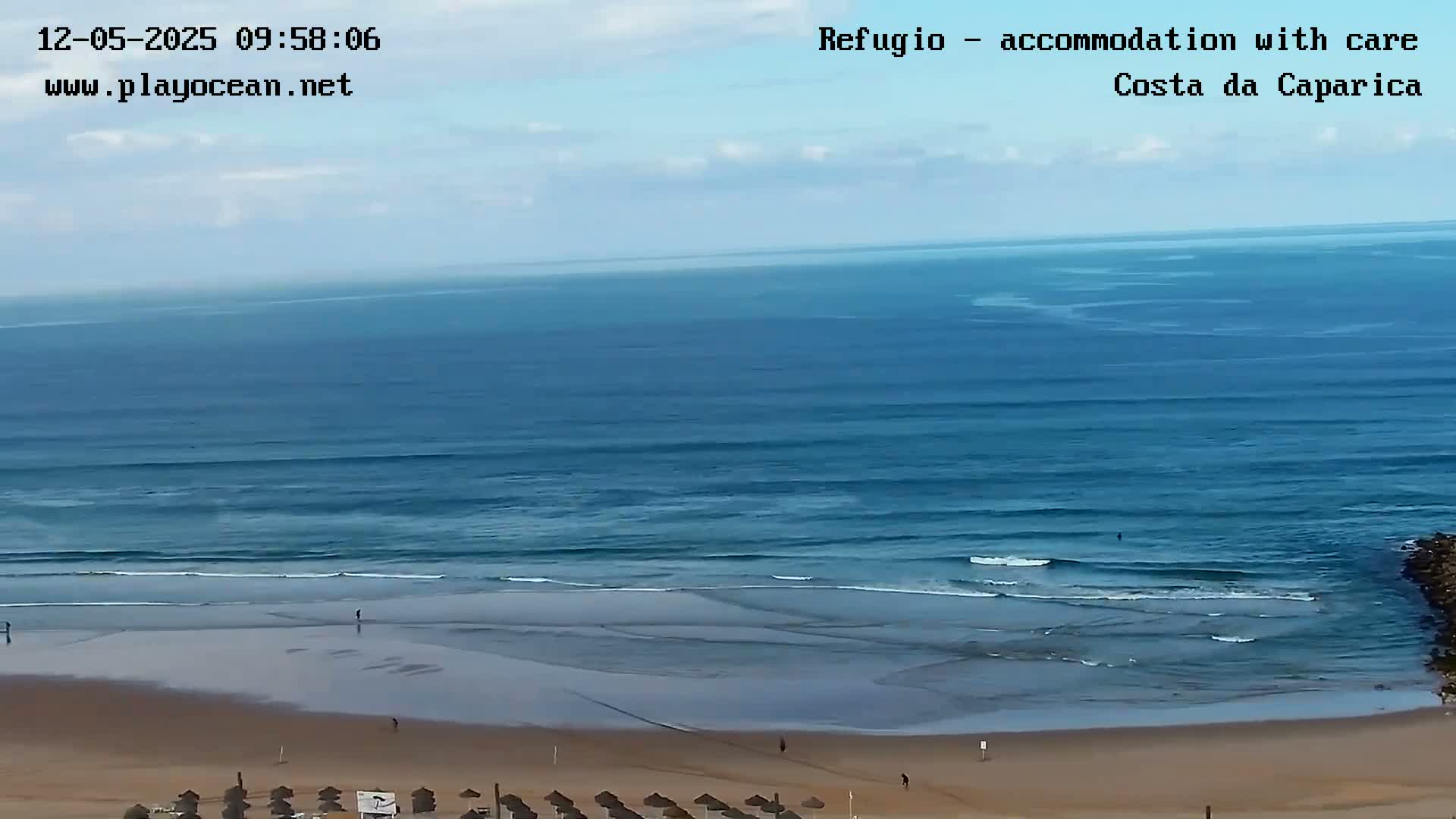 A mostly sunny day shows a wide expanse of ocean with gentle waves rolling onto a sandy beach, where a few people are walking and some beach umbrellas are set up.