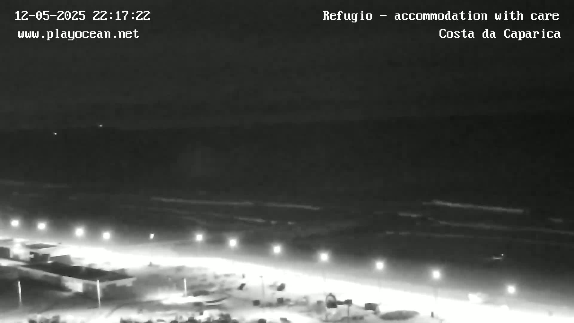 A nighttime, low-light view of a beach with dimly lit structures and a dark ocean in the background.