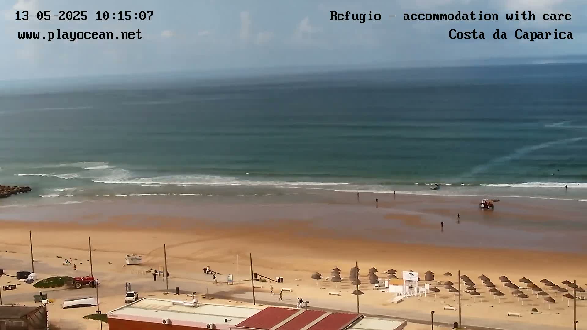 A sandy beach with calm ocean waves, beach umbrellas, and a few people is viewed from an elevated vantage point on a partly cloudy day.