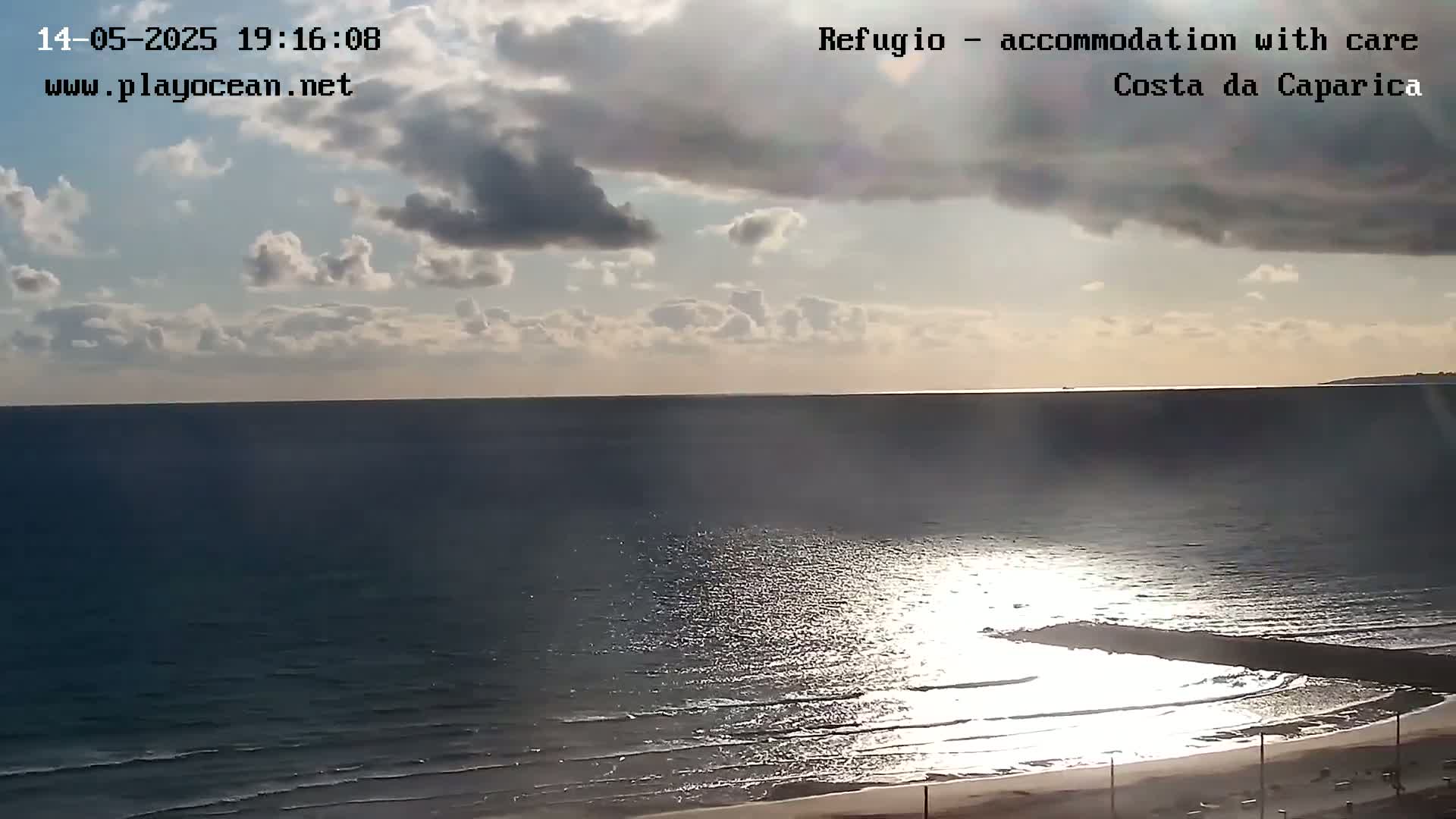 A mostly cloudy afternoon shows the ocean meeting a sandy beach, with sunlight glinting on the water.