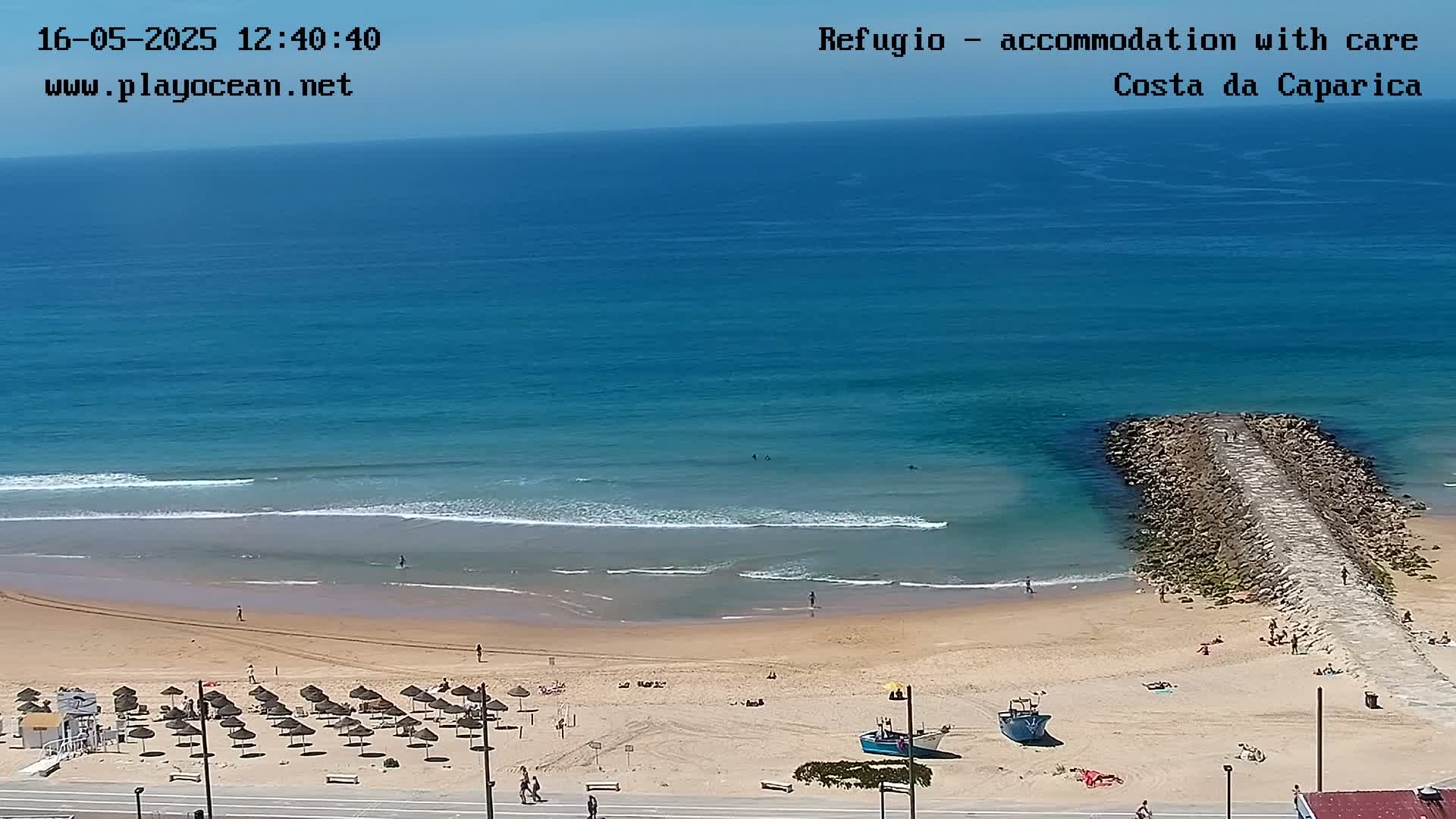 A sandy beach with calm ocean waves, numerous beach umbrellas, and a rock jetty extends into the water under a clear, sunny sky.