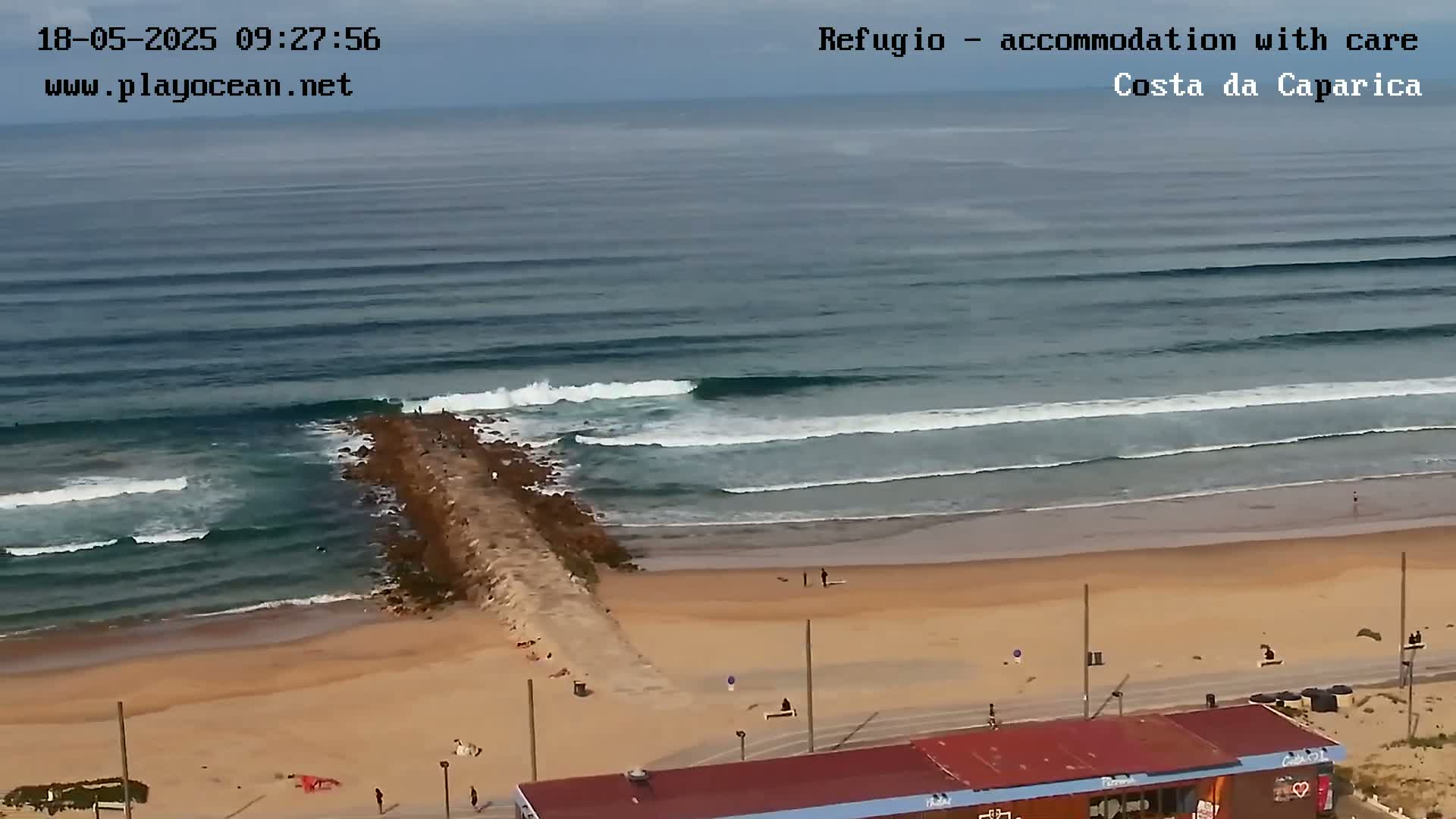 An aerial view shows a sandy beach with a long rock jetty extending into the ocean, where waves are rolling in under a mostly cloudy sky.