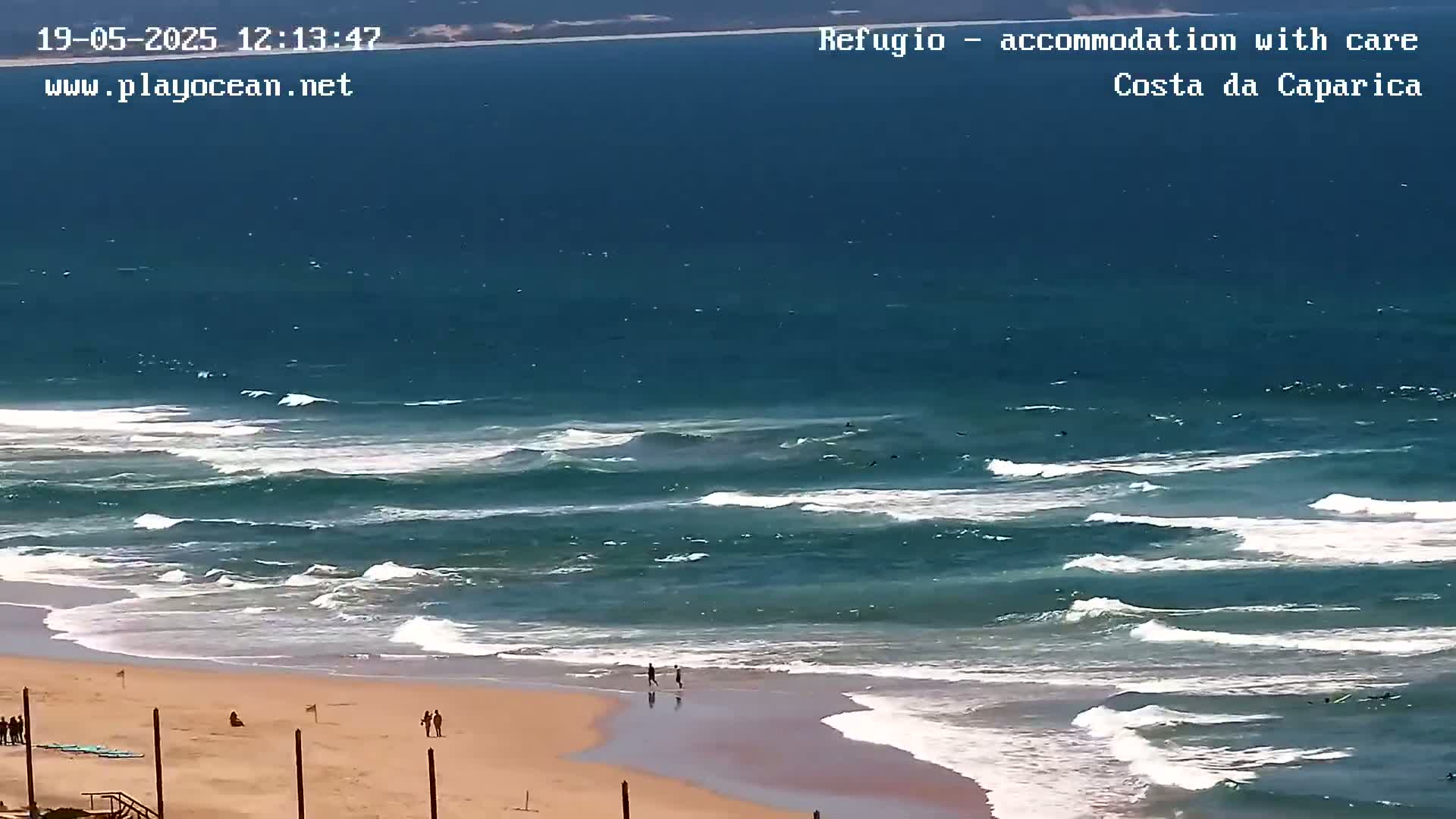 A sandy beach with a few people on it is bordered by a dark-blue ocean with moderate waves under a bright, sunny sky.