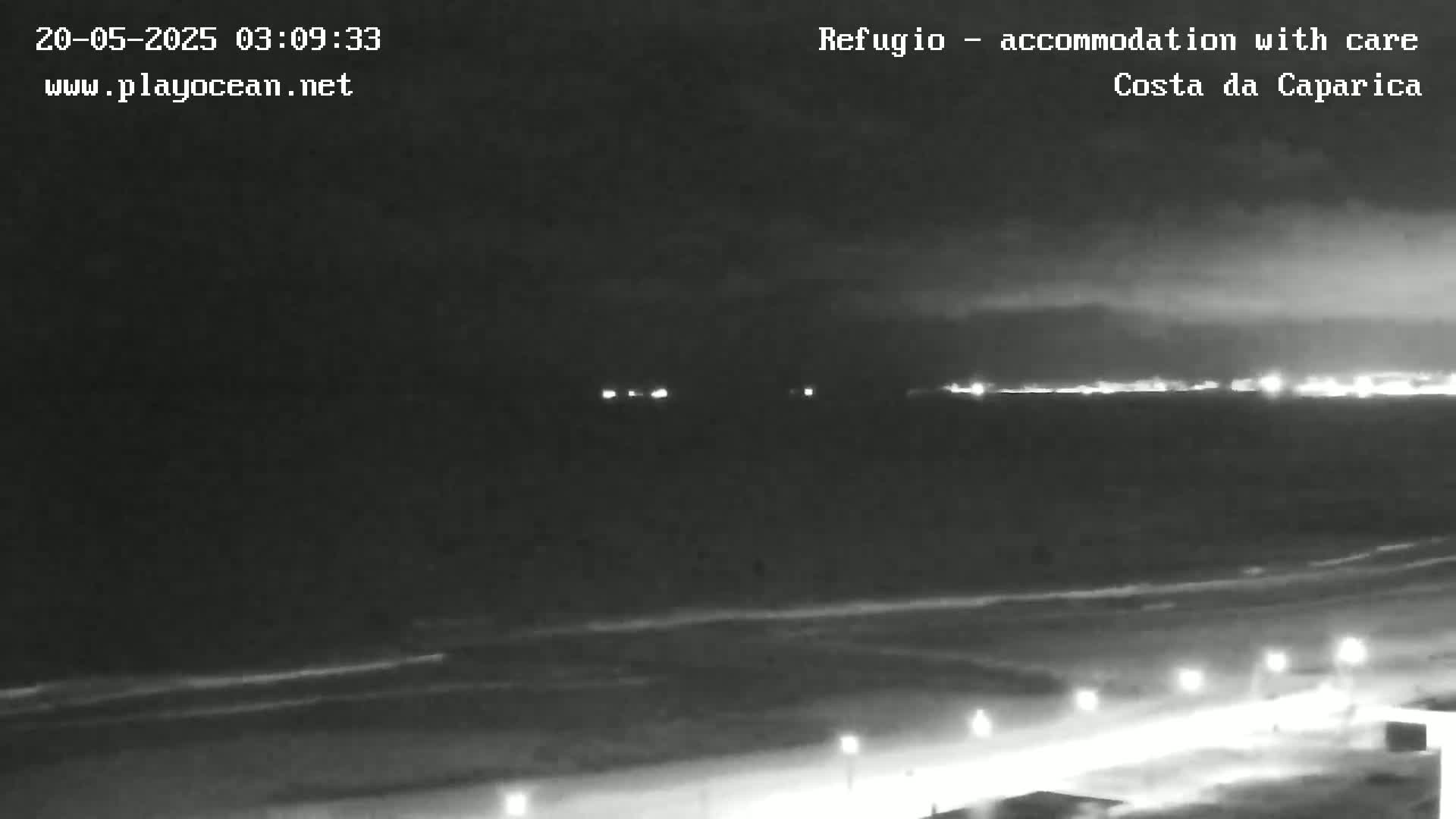 A nighttime view of a dark ocean beach with a distant city skyline faintly illuminated under a cloudy sky.