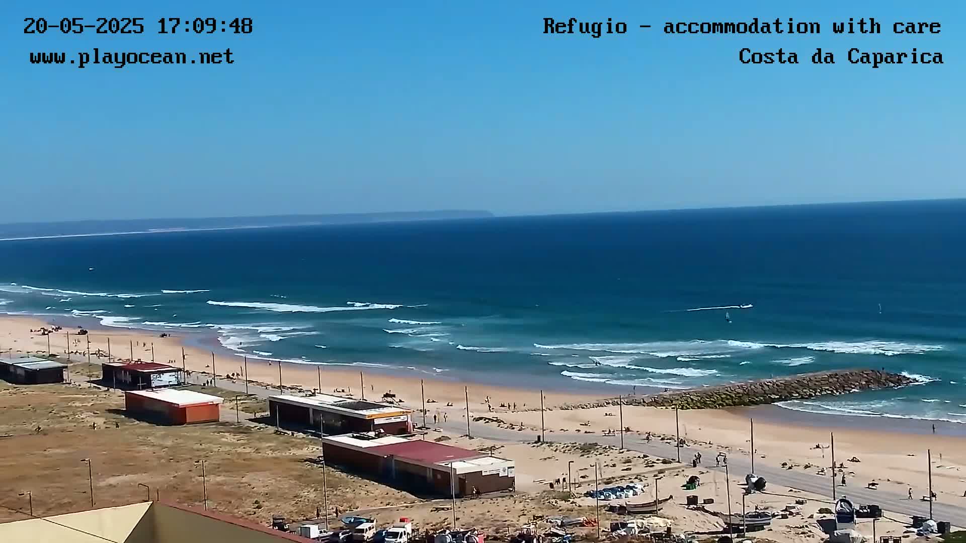 A beach with small waves under a clear blue sky, showing a sandy shore with buildings and people, and a distant landmass on the horizon.