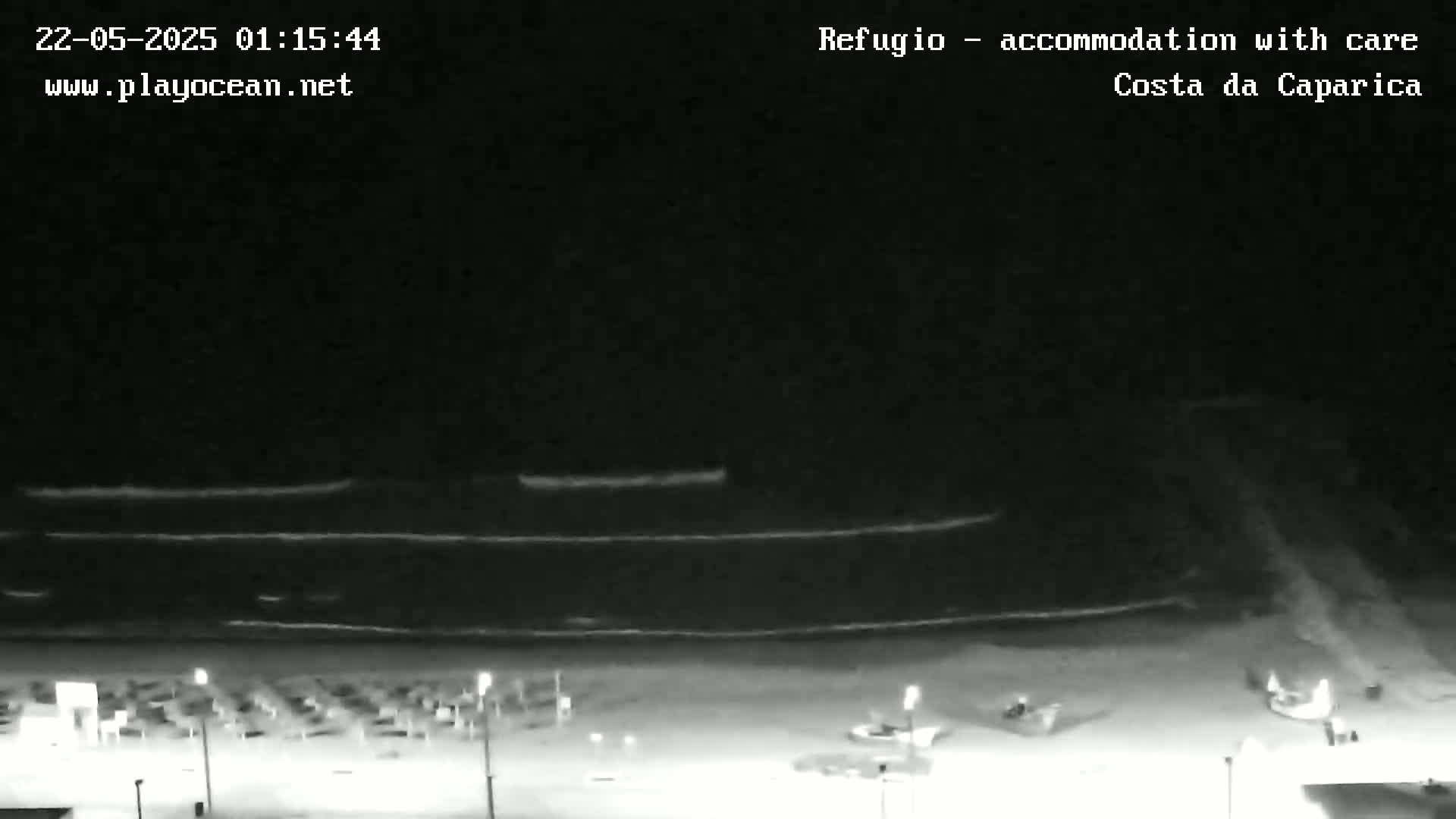 A nighttime beach scene shows dark water with small waves, a dark sandy shore with sparsely placed furniture, and several indistinct figures near the water's edge.