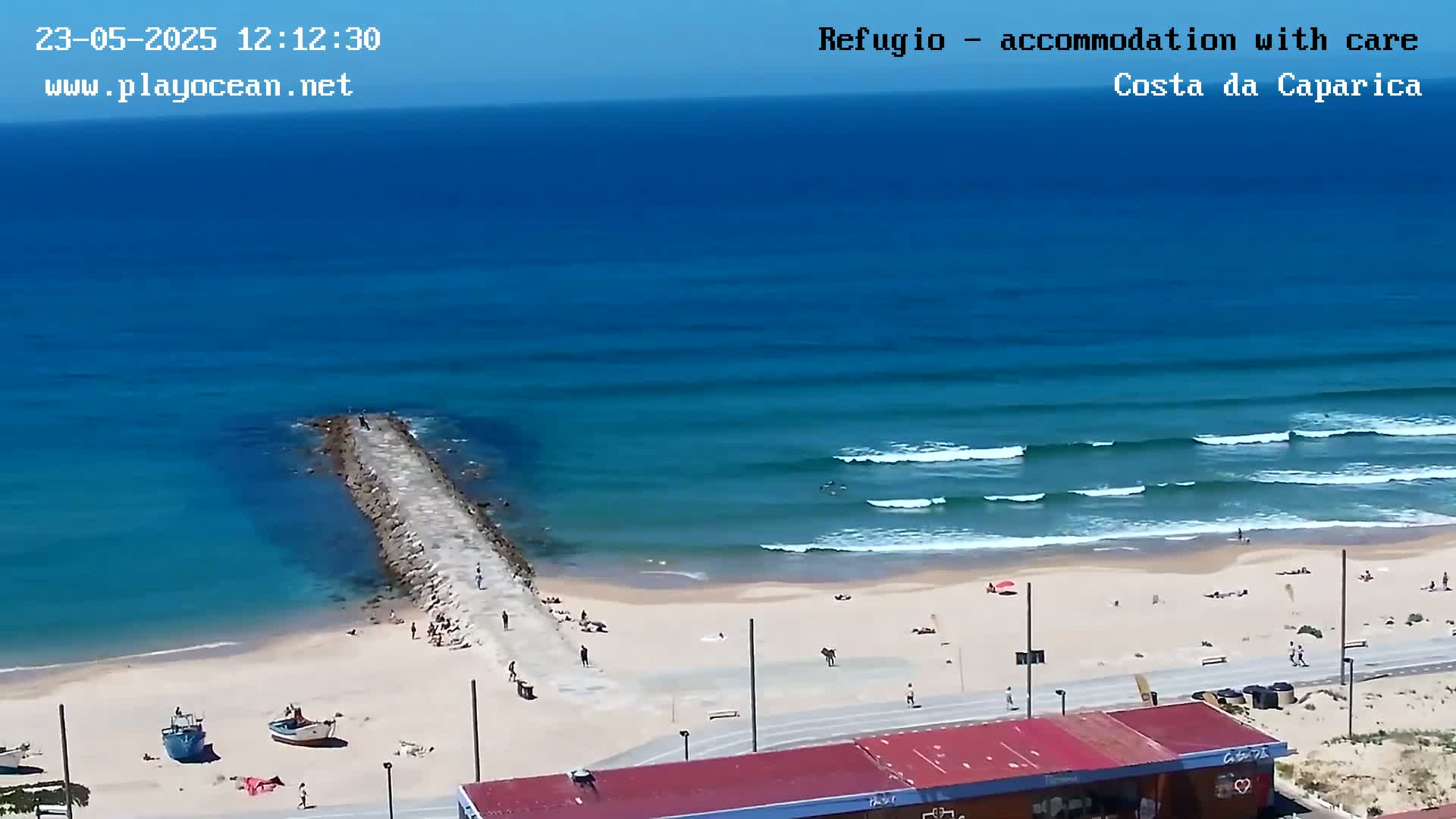 An overhead view shows a sandy beach with a long jetty extending into a blue ocean with small waves under a sunny sky.