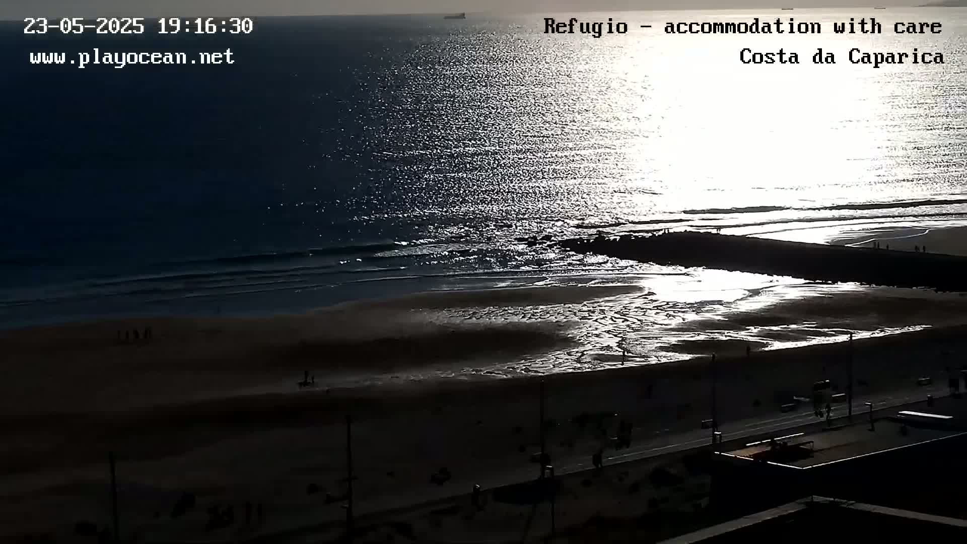 A sandy beach with a calm ocean reflecting sunlight at sunset, viewed from above.