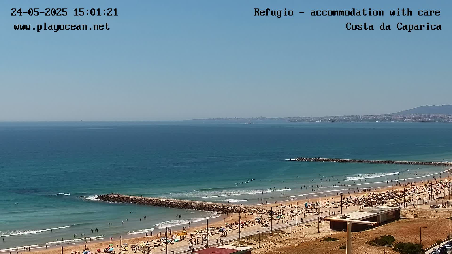 A sunny day at a crowded beach with a calm ocean, featuring a long, rocky breakwater extending into the water.