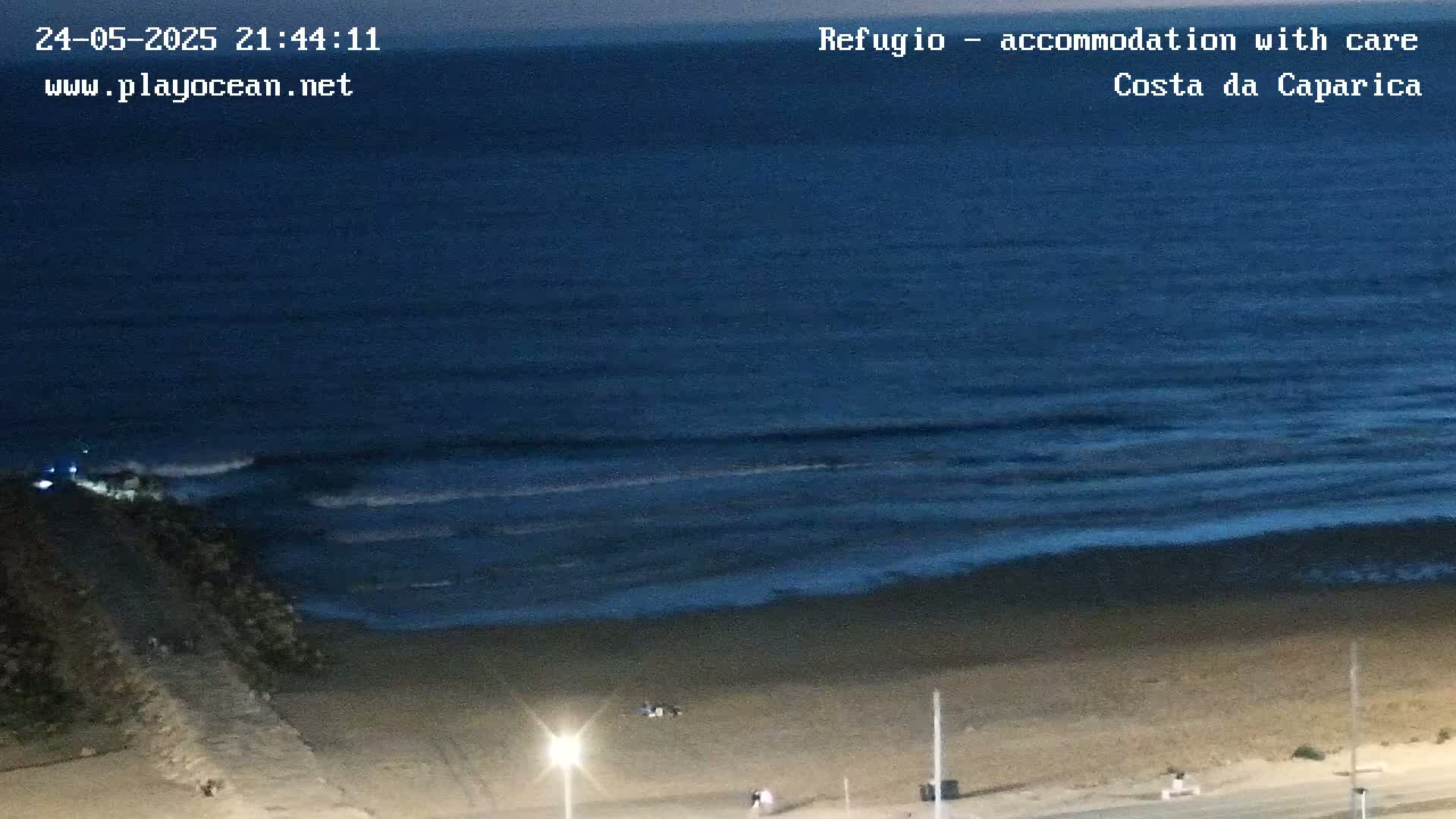A dark blue ocean with small waves rolls onto a sandy beach at night, with a few people visible on the sand.