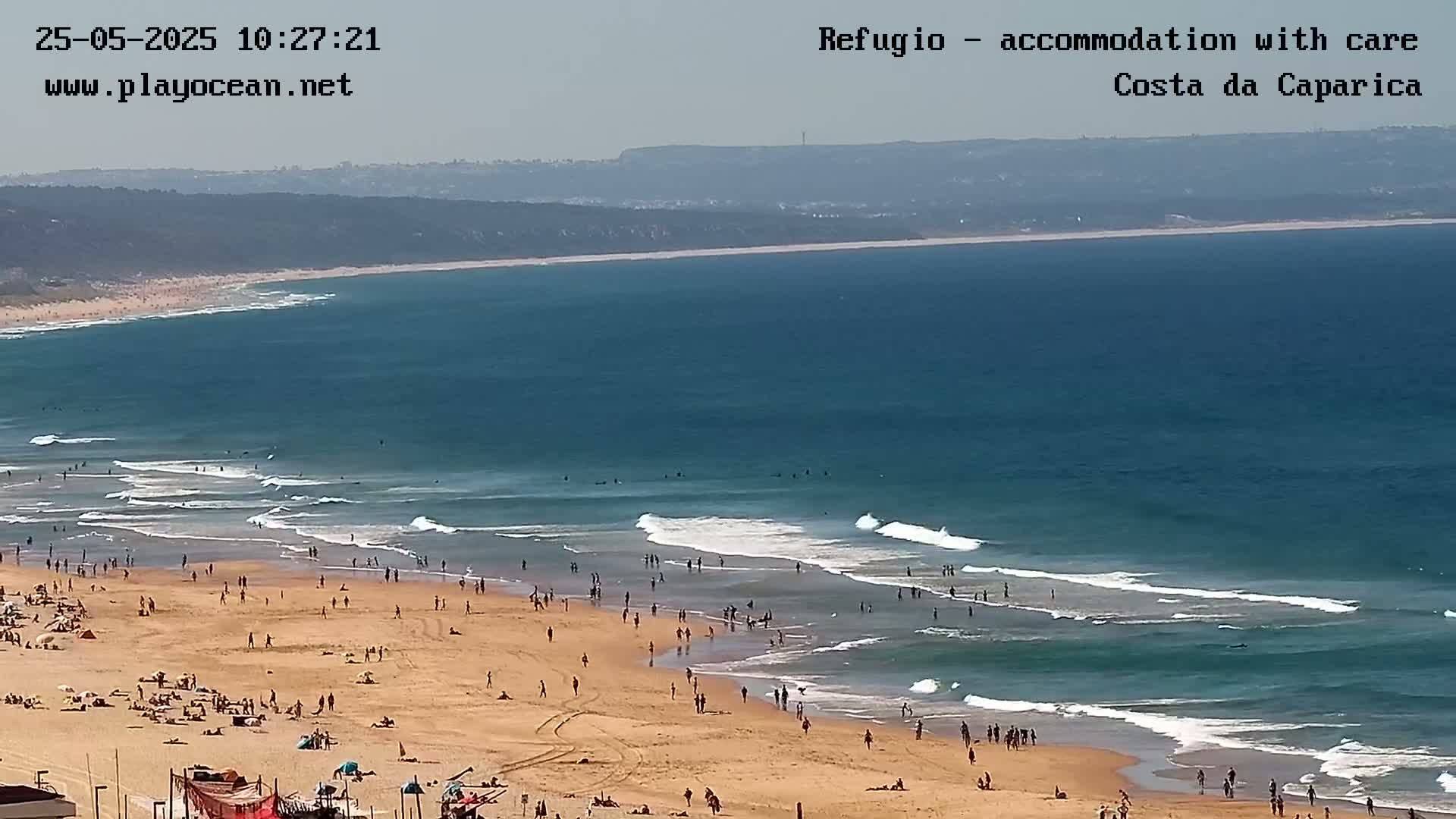 A sandy beach with many people on it is bordered by a blue ocean with small waves on a hazy day.