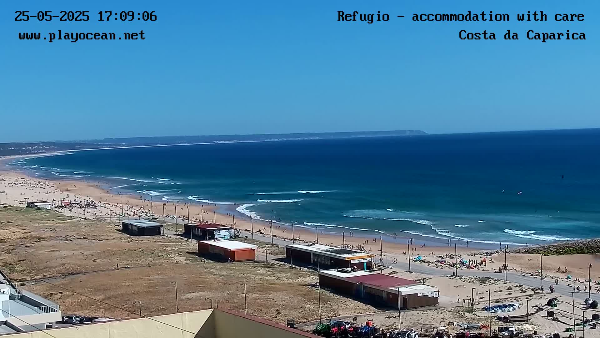A long sandy beach with many people on it is seen from a high vantage point on a sunny day, with moderate waves breaking in the ocean.